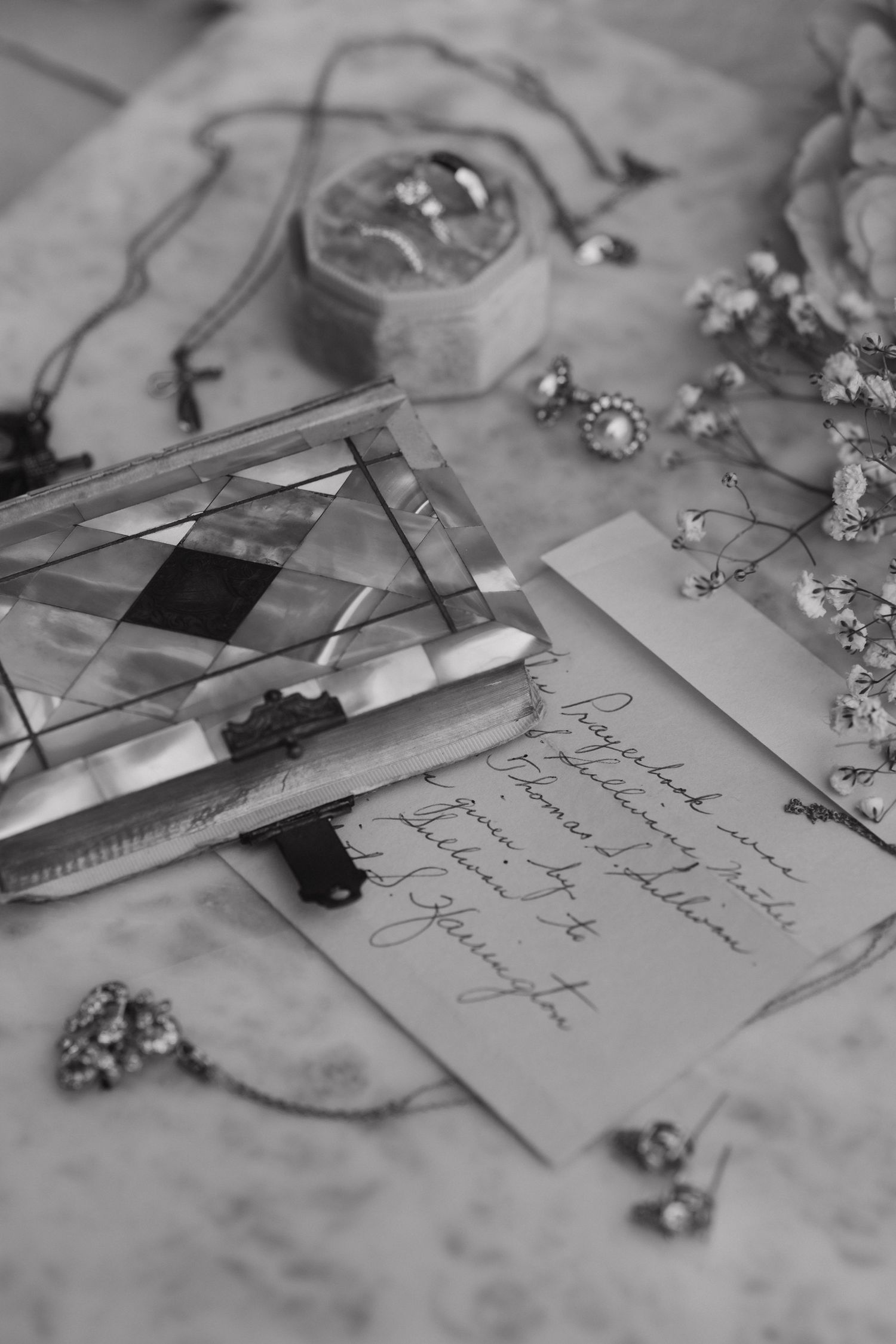 Black and white flat lay of wedding stationery, jewelry, dried flowers, and a geometric glass trinket box on marble.