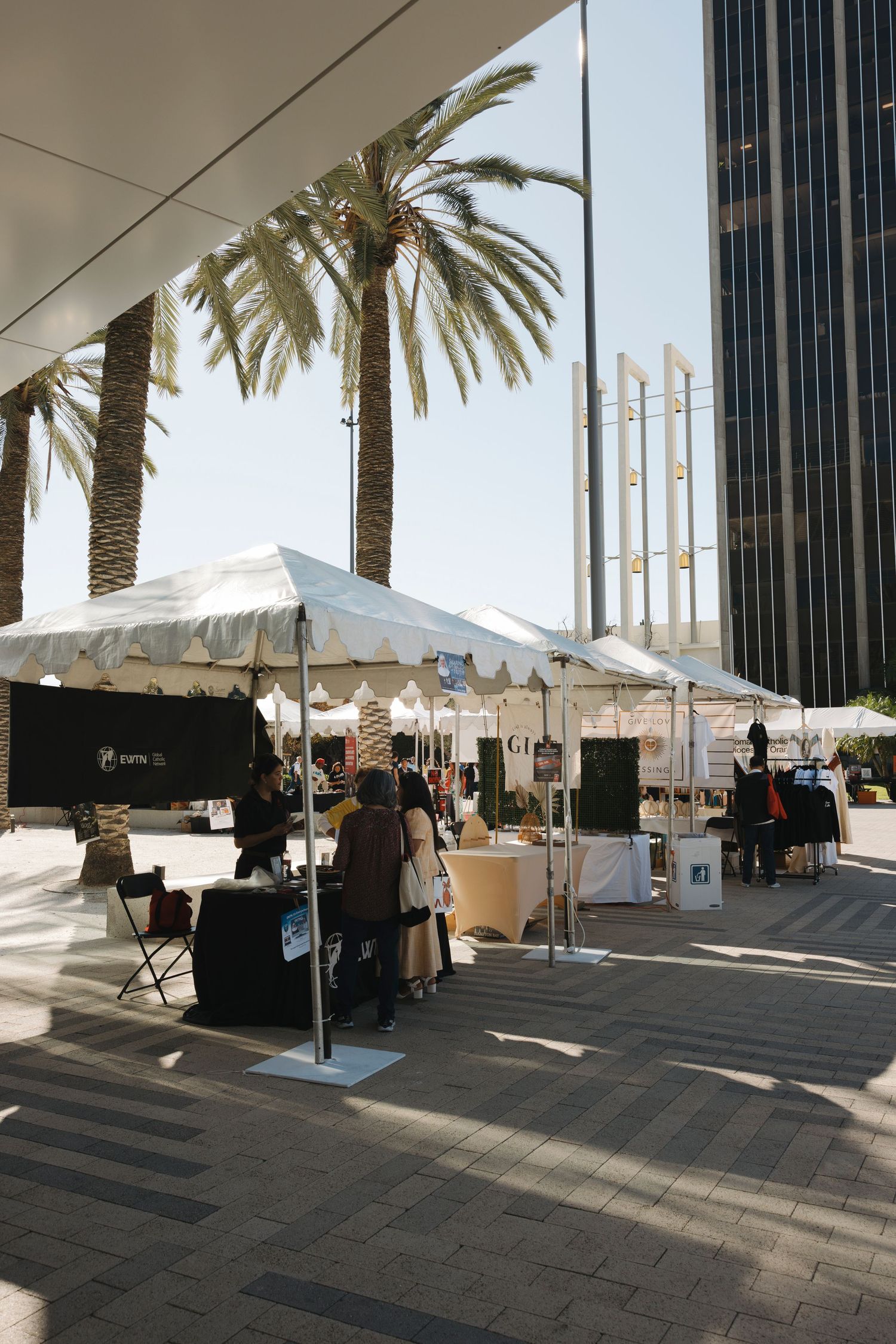 White event tents and vendor booths line a plaza with palm trees and modern buildings.