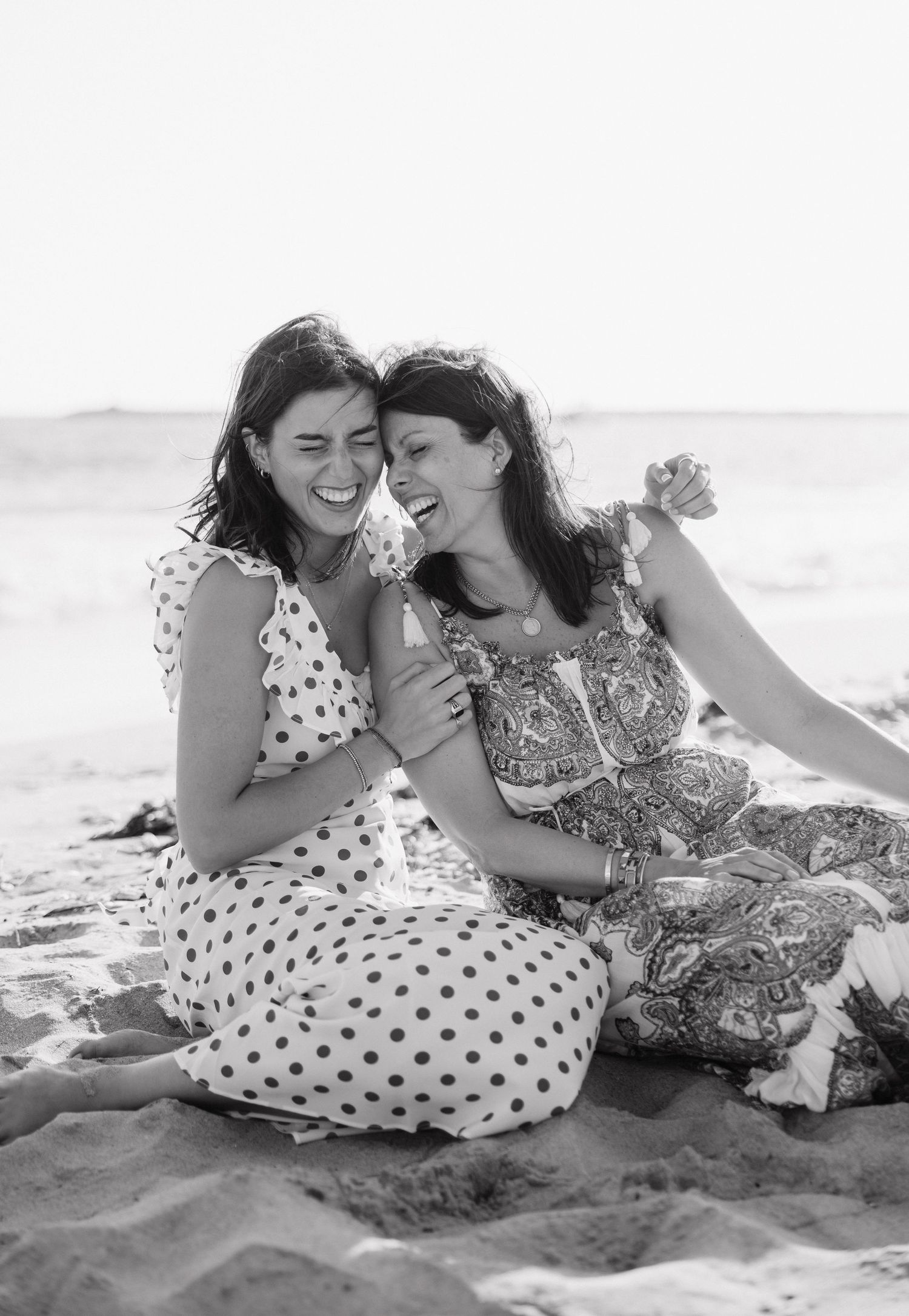 Two women laughing together on a beach, wearing summer dresses, sitting on sand with ocean in background.