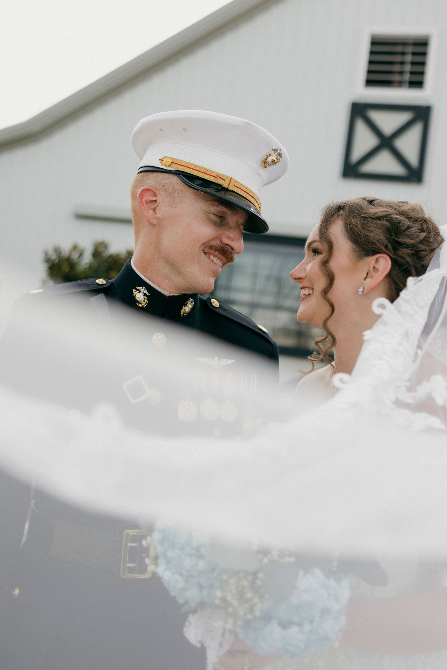 A smiling military groom in dress uniform gazes at his bride, her veil billowing in the foreground near a white barn.