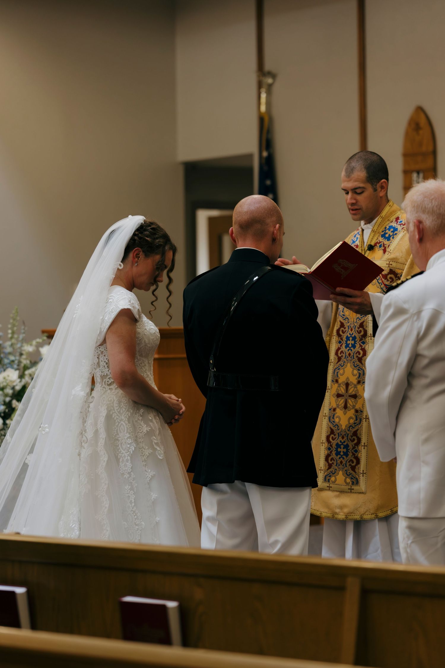 Bride and groom exchange vows at church altar with officiant reading from book during wedding ceremony.