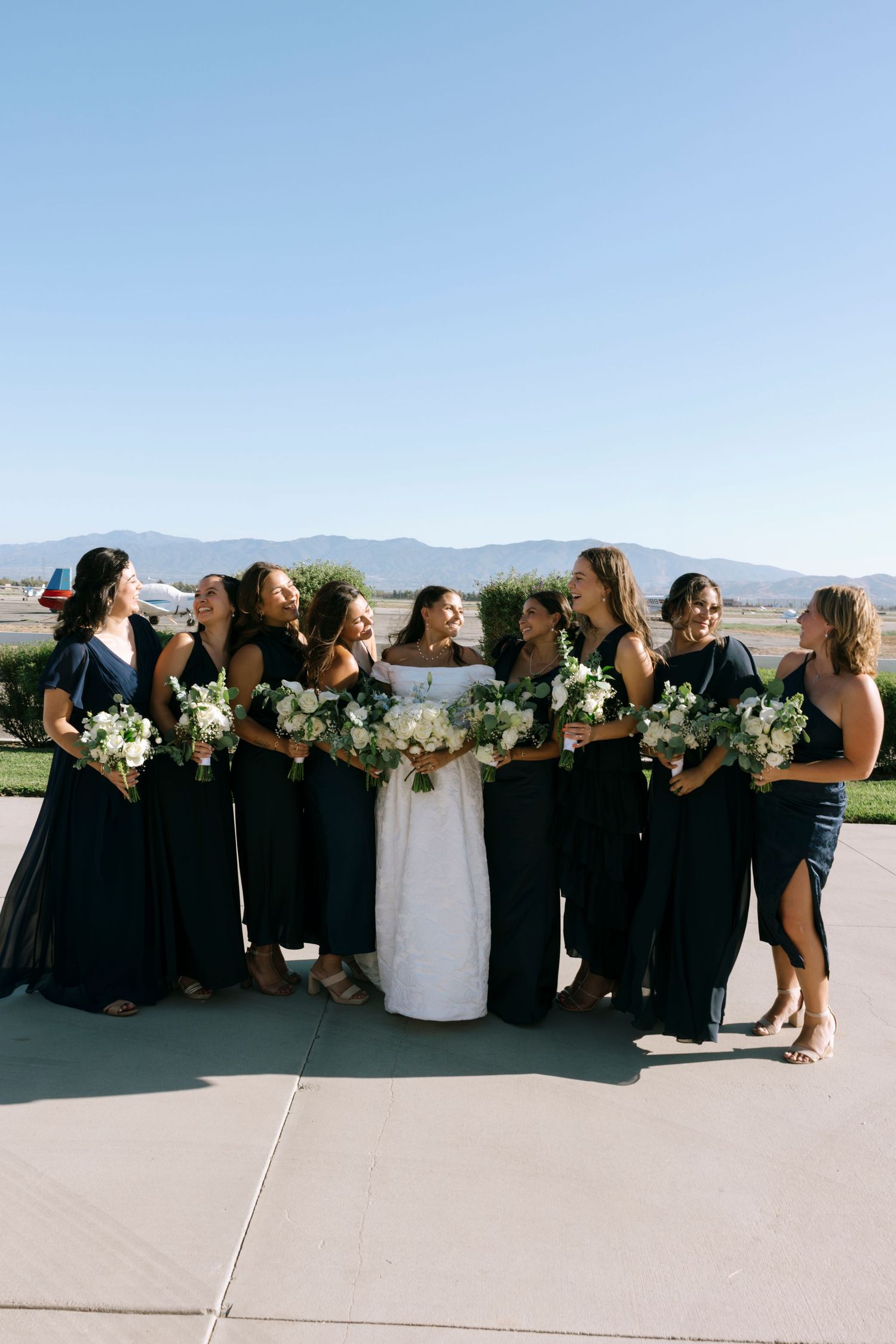 Wedding party in black dresses and white gown arranged in curved line outdoors.