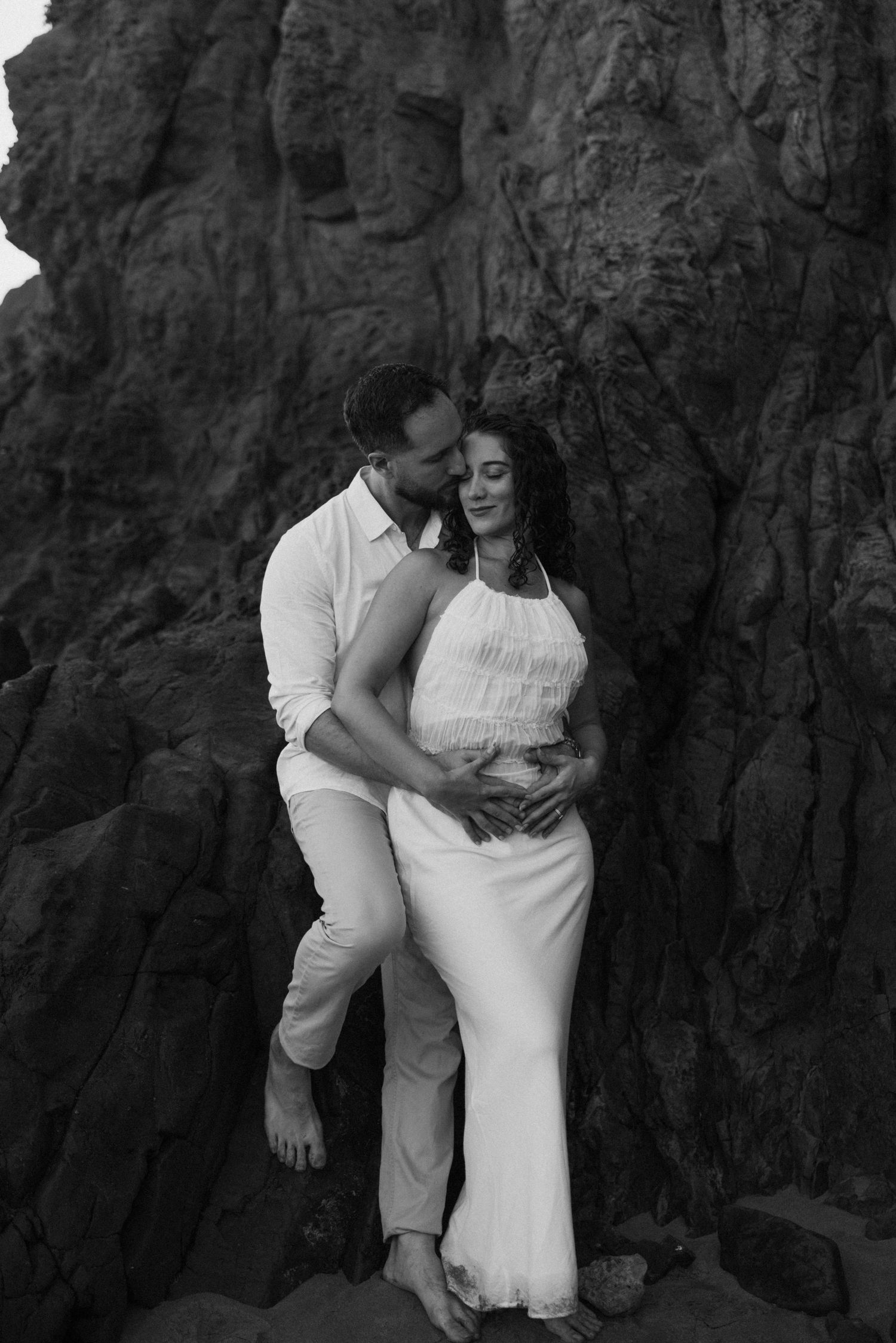 Couple embracing against dramatic rocky cliff backdrop in black and white.
