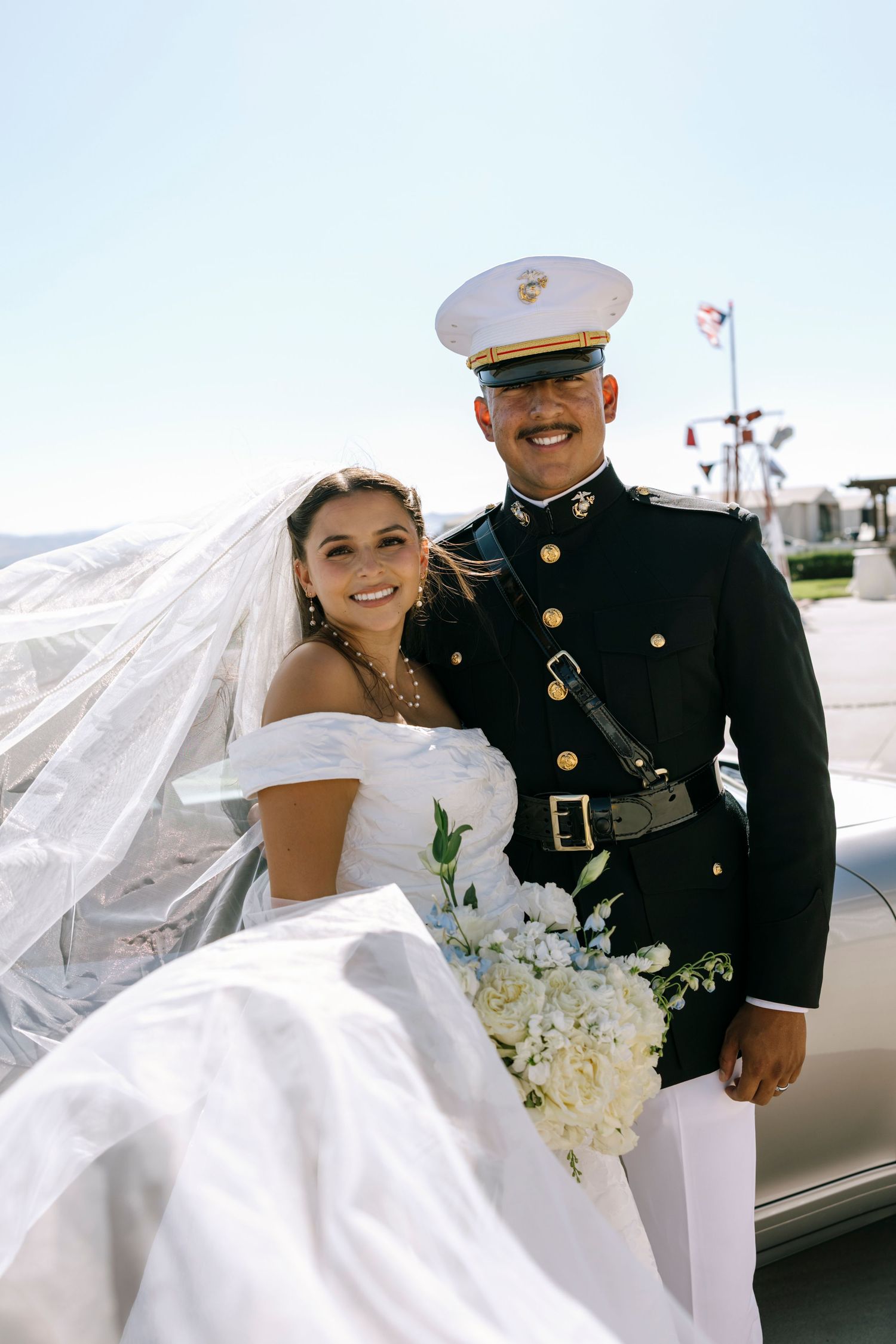Military wedding couple shares romantic moment with flowing veil against bright sky.