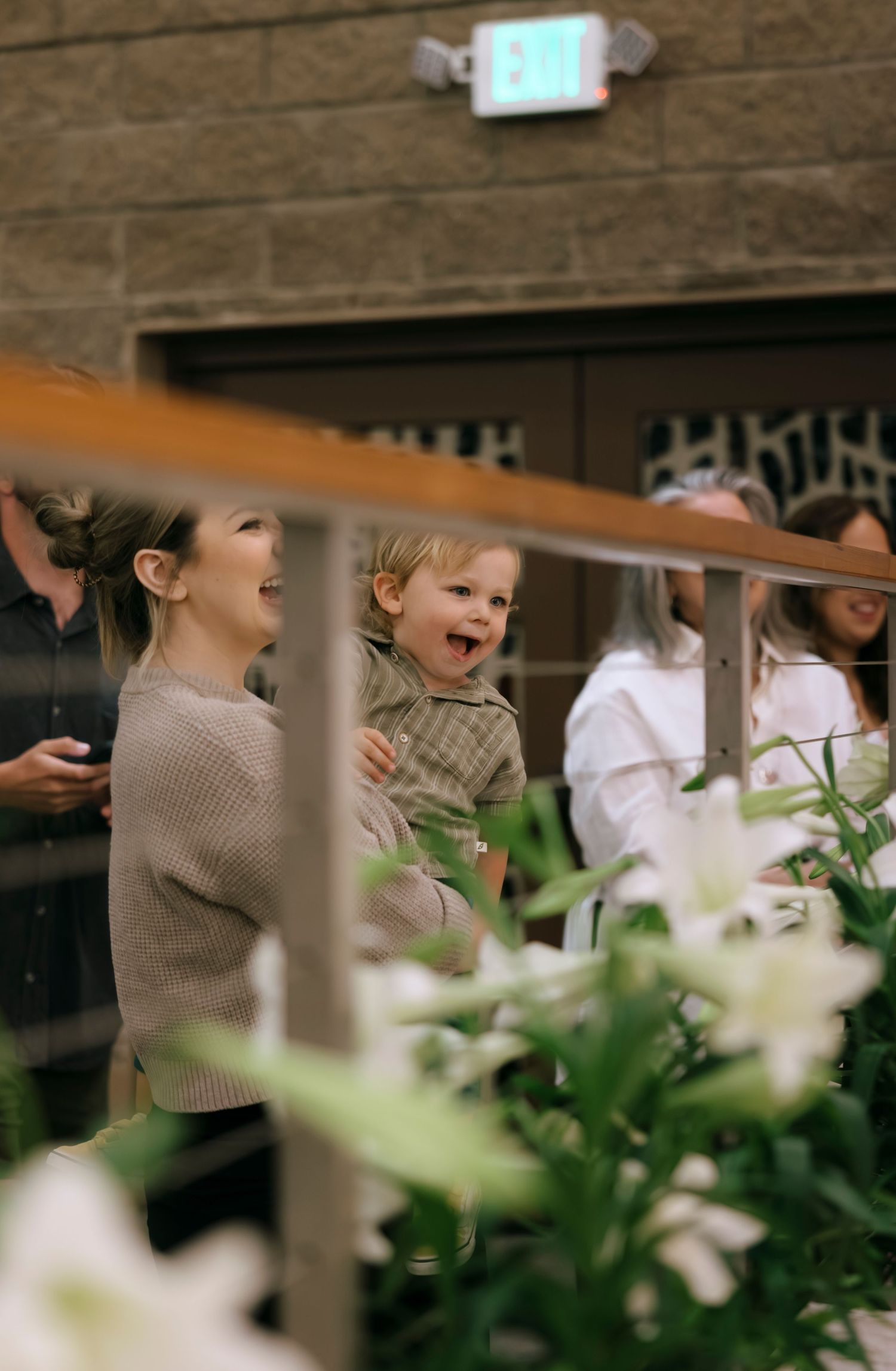 Smiling mother holding a laughing toddler while watching a church ceremony near white lily flowers.
