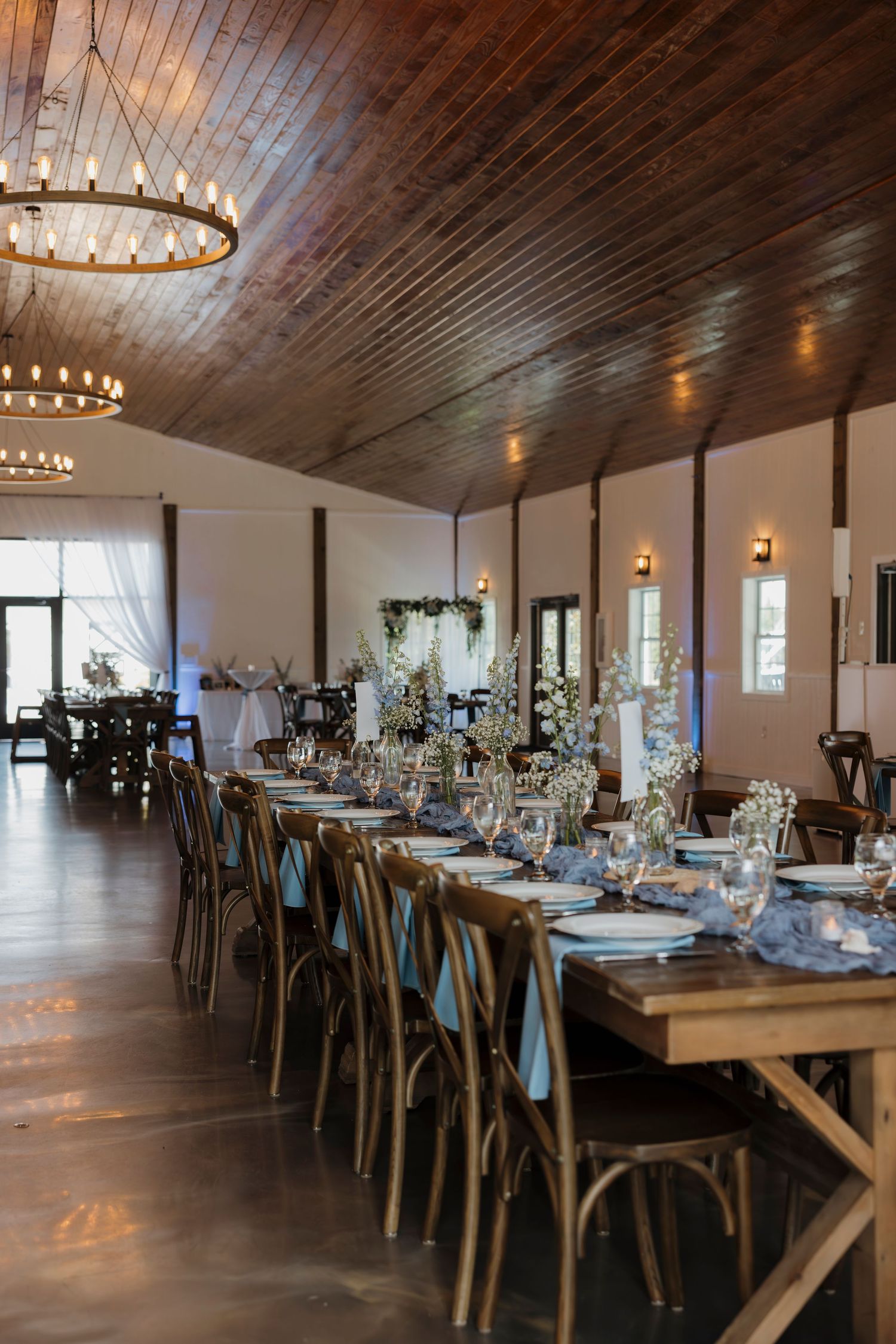 Elegant wedding reception hall with long farm tables, cross-back chairs, floral centerpieces, and warm wood ceiling lighting.