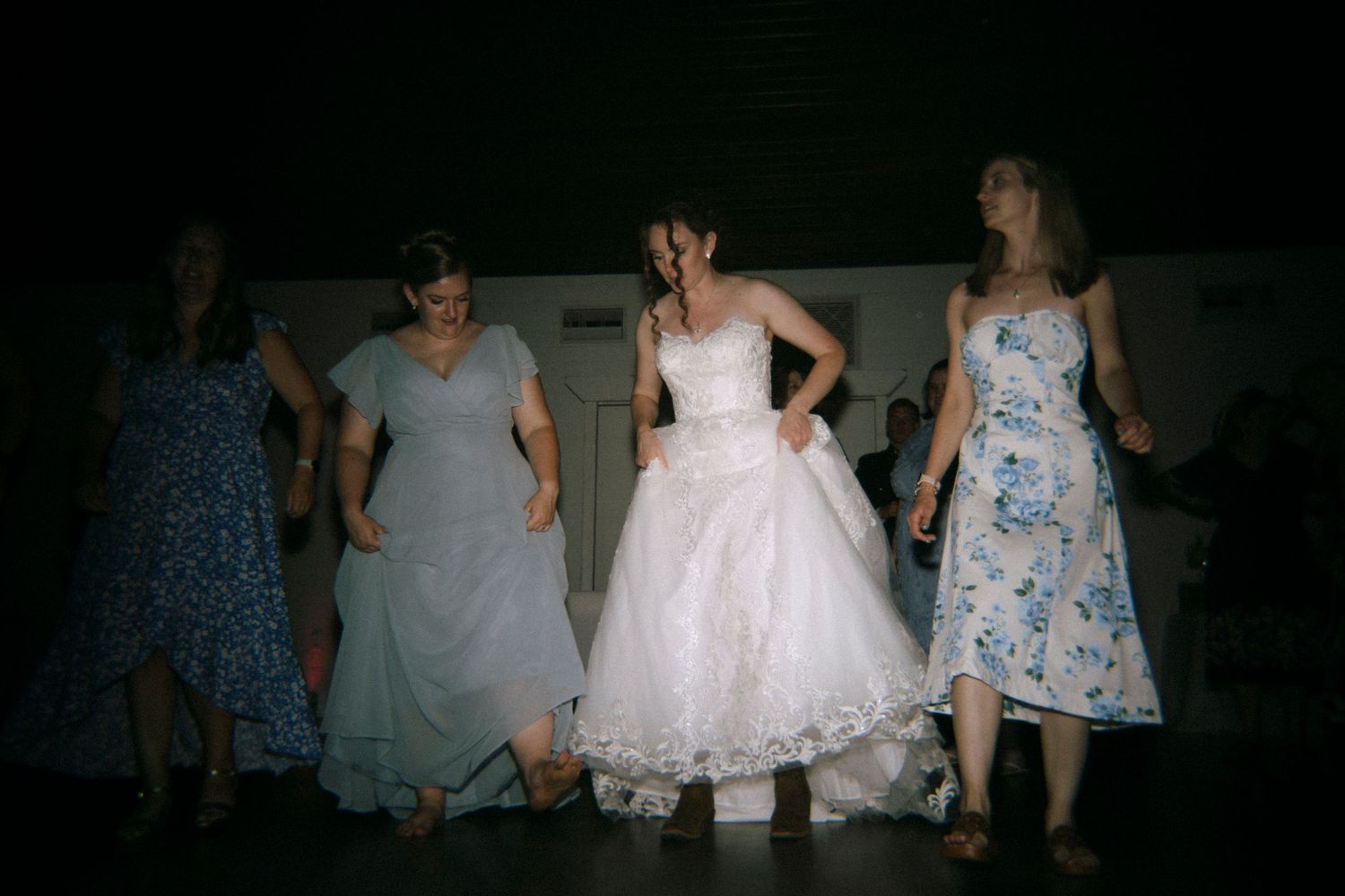 Bride in white gown dancing with bridesmaids in floral dresses at a wedding reception.