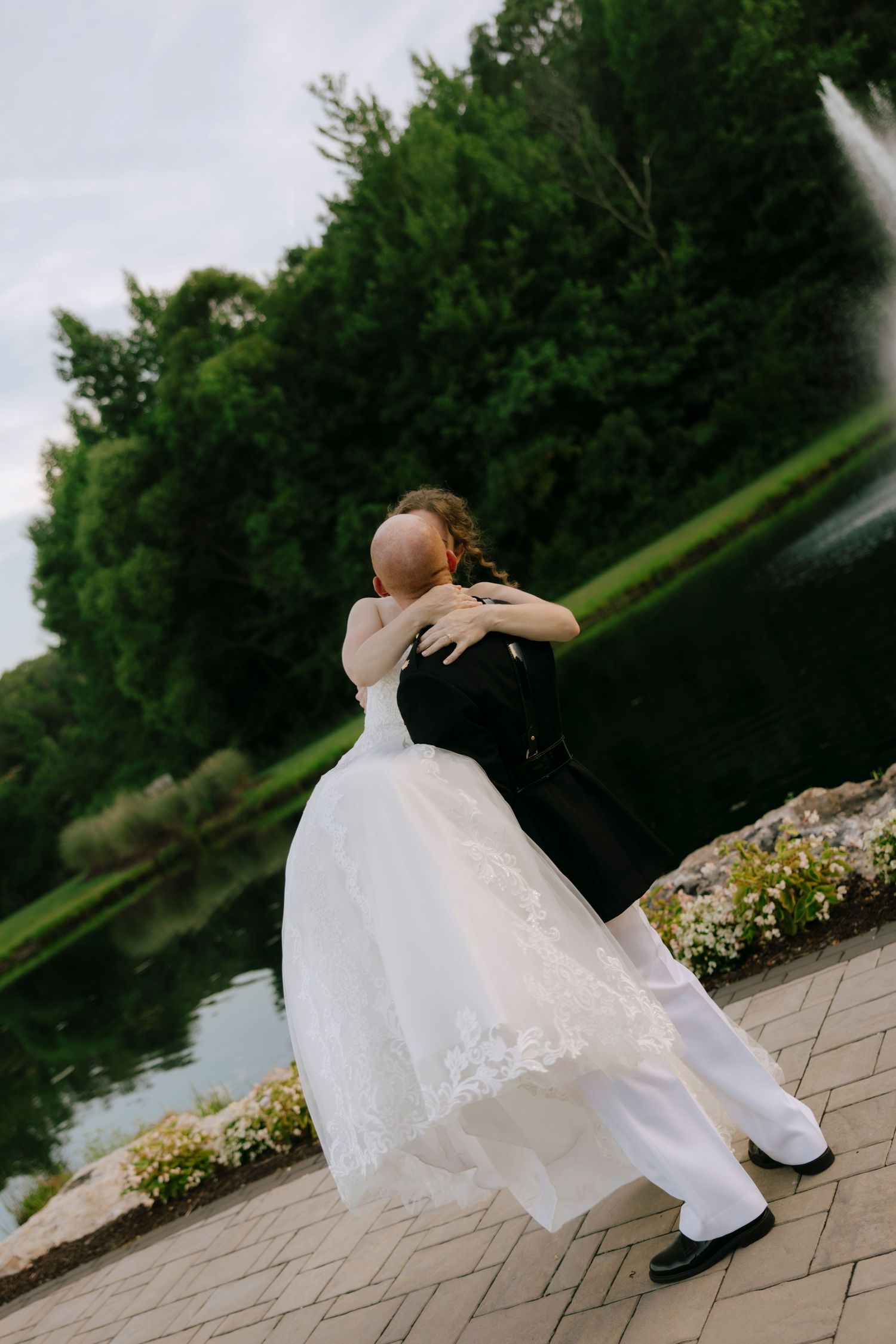Bride and groom embracing outdoors near a pond, surrounded by lush green trees on their wedding day.