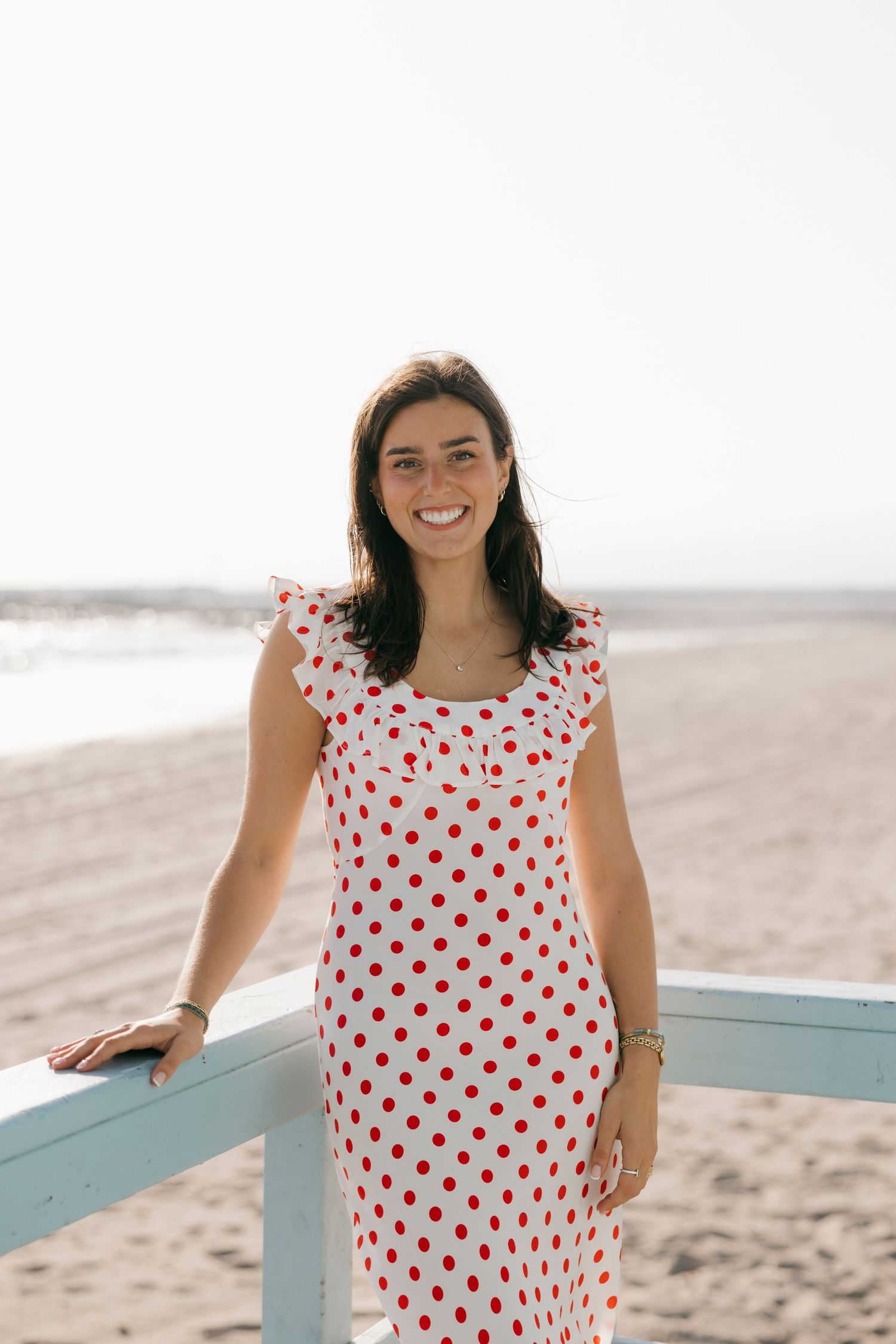 Smiling woman in white red polka dot dress standing by beach railing on sunny day.