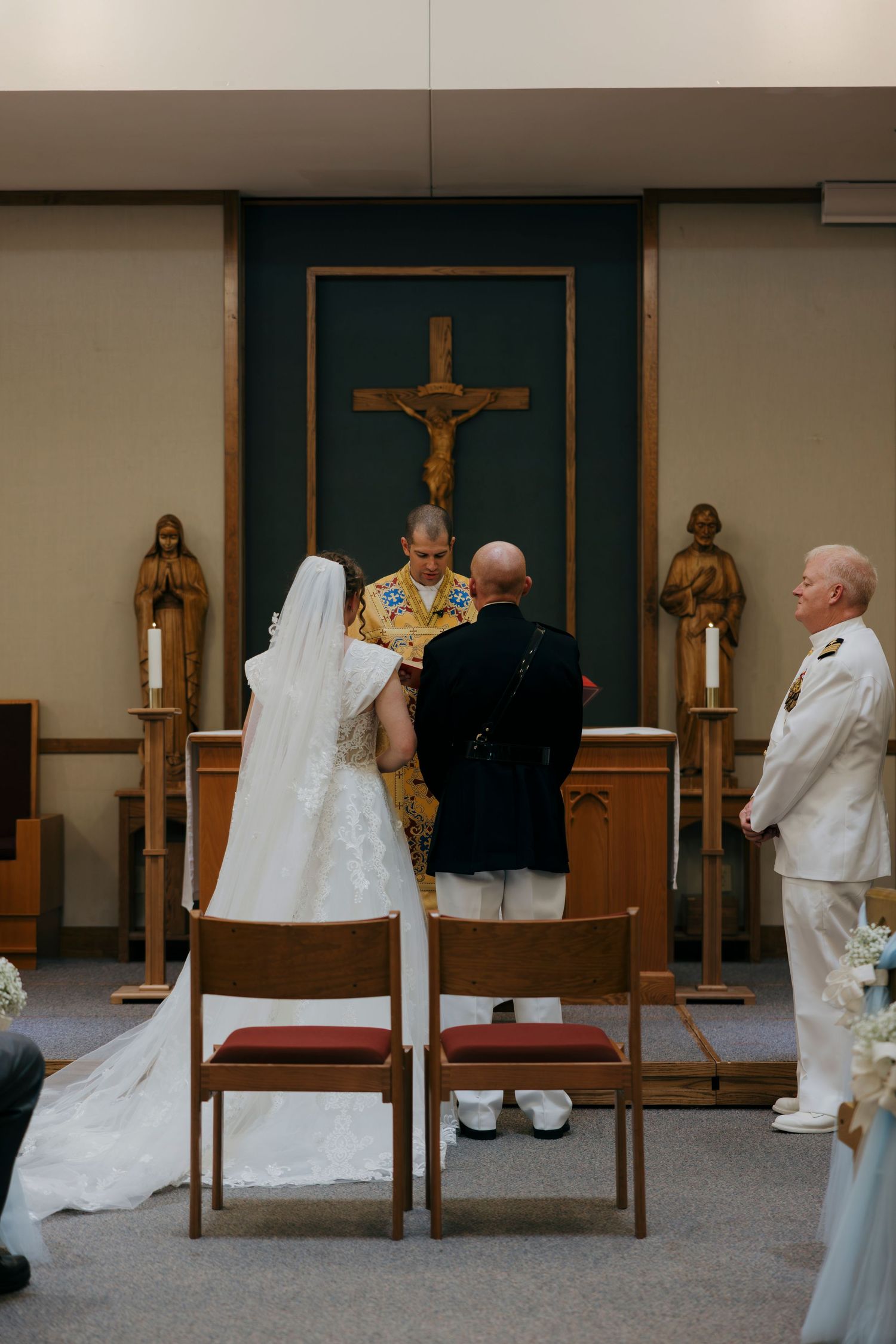 Bride and groom exchange vows at church altar before officiant, with crucifix backdrop and candles, as guest in white uniform watches.