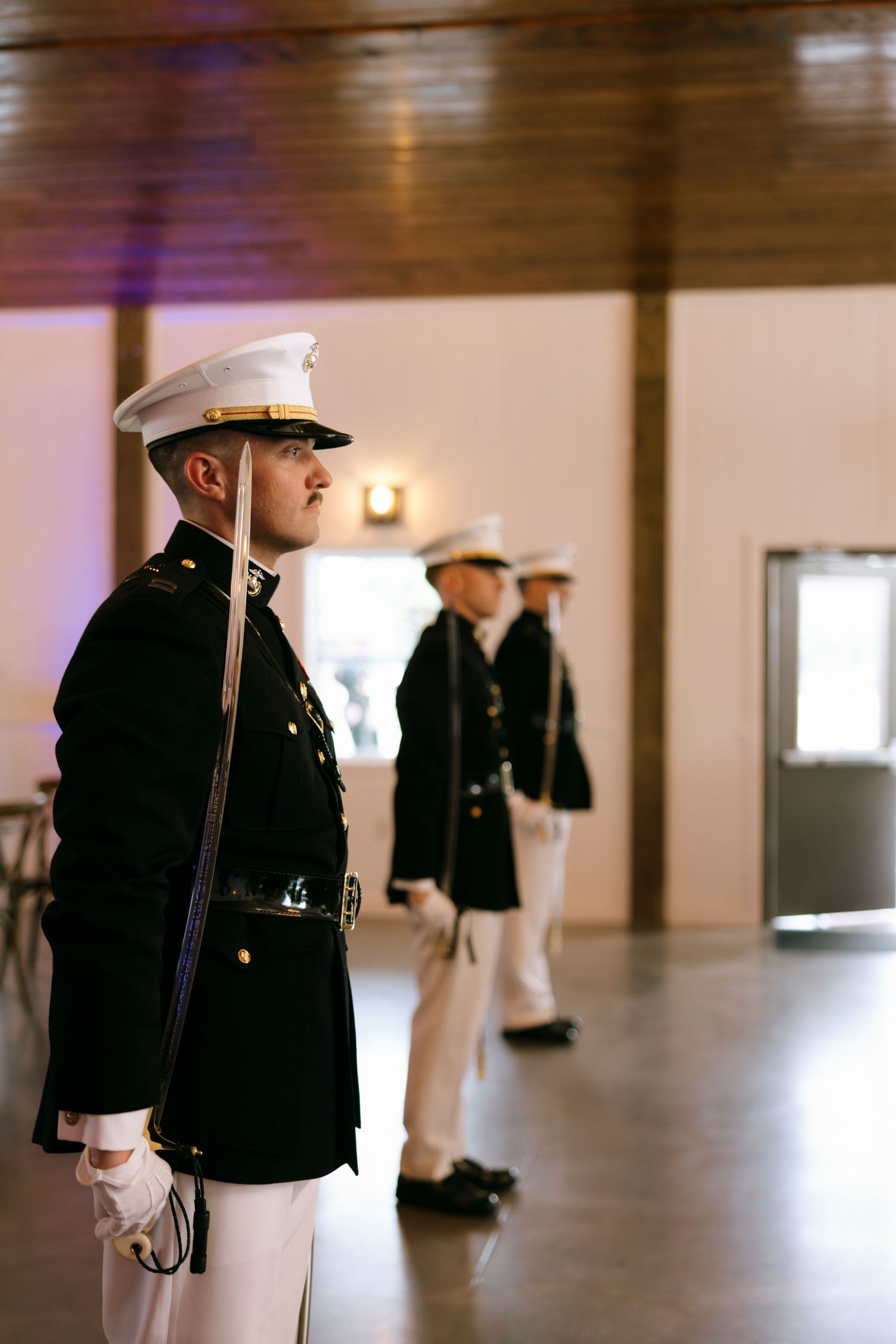 Military officers in dress uniforms stand at attention inside a formal venue during a ceremony.