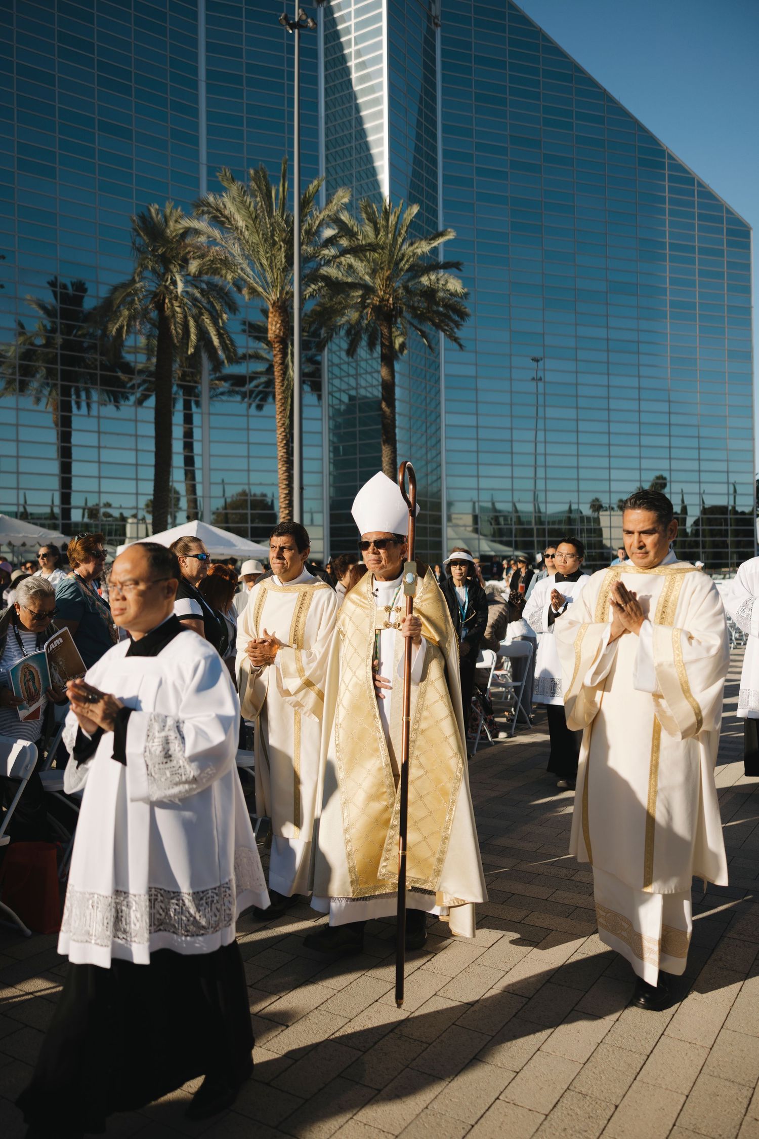 Clergy members in white and gold vestments process outside a modern glass building.
