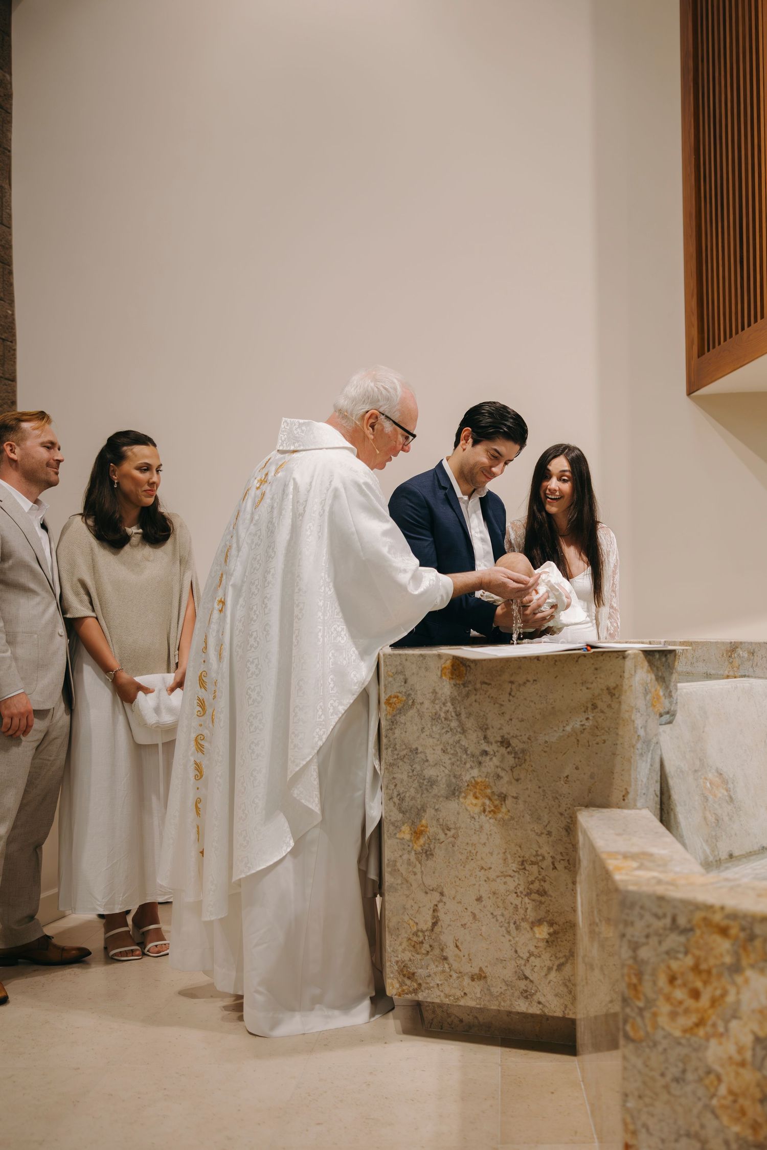 Catholic priest performing infant baptism at marble font with parents and godparents present in modern church.