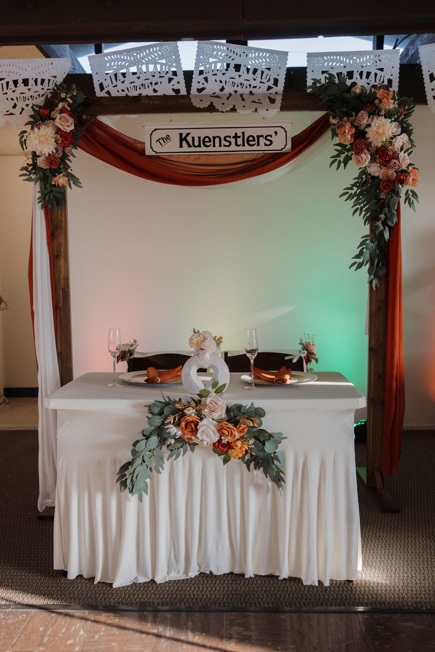 Wedding reception head table decorated with white linens, floral arrangements and Kuenstlers signage.