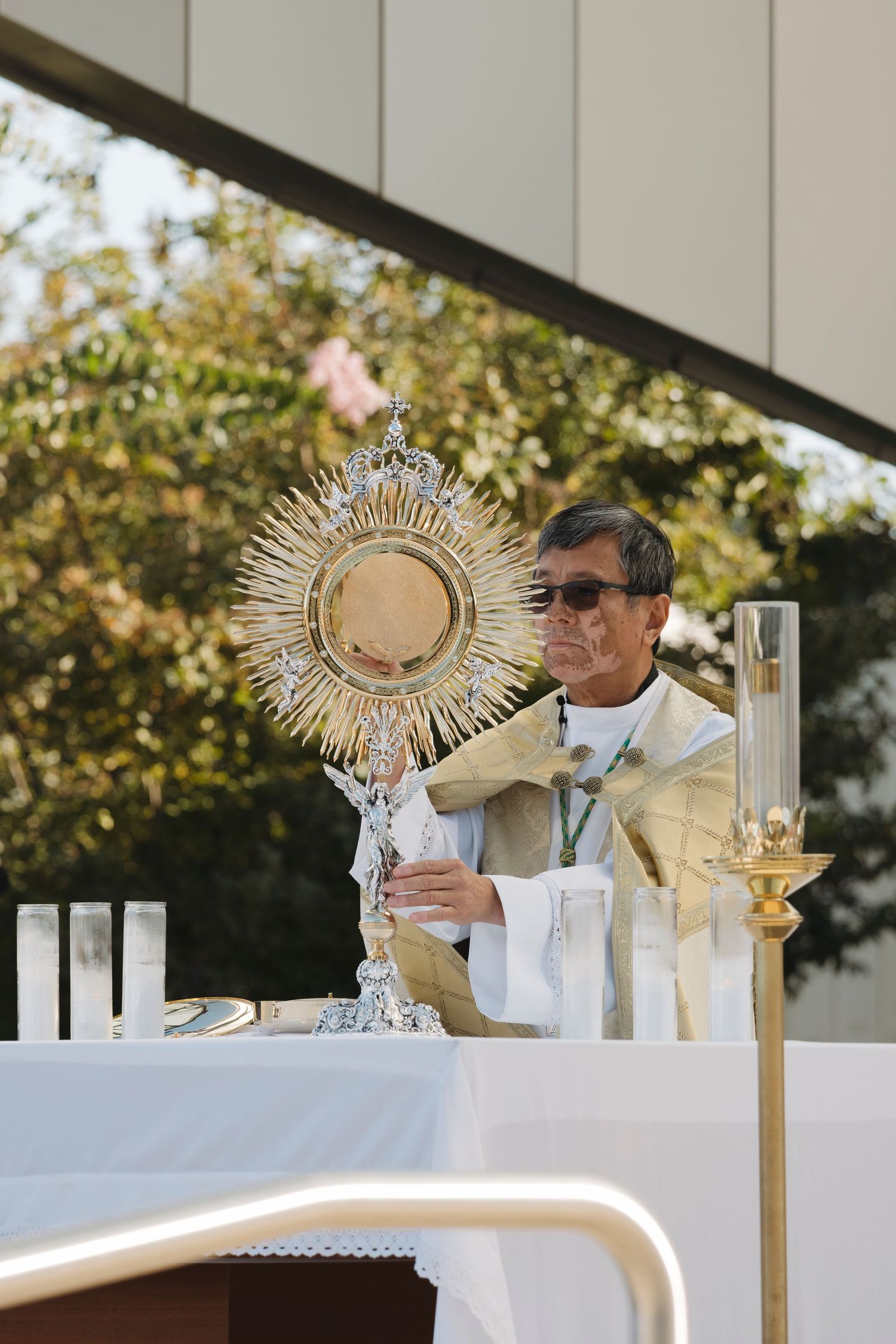 Priest holds up ornate golden monstrance during Eucharistic adoration at outdoor altar.