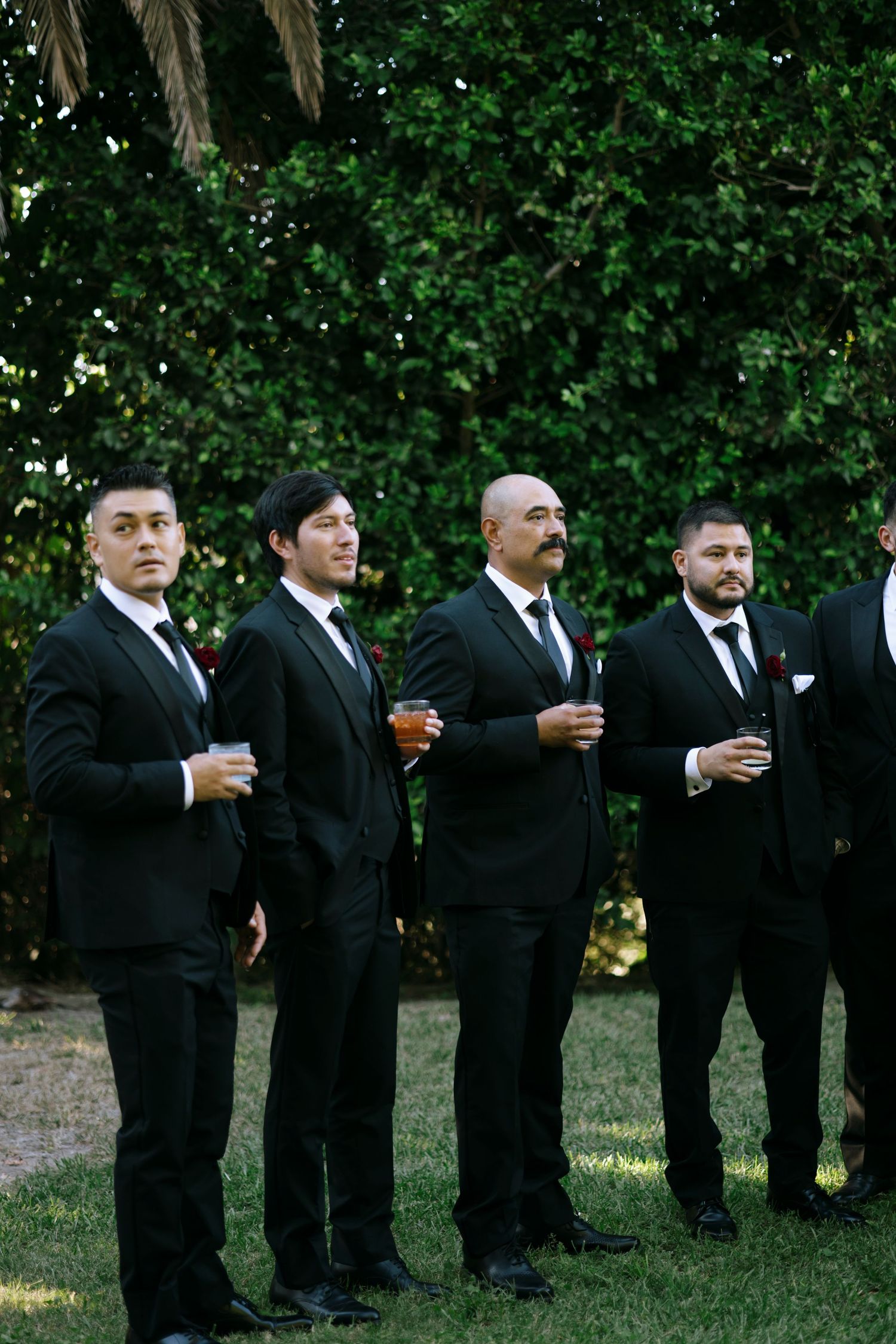 Wedding party members in formal black suits holding drinks during outdoor celebration.