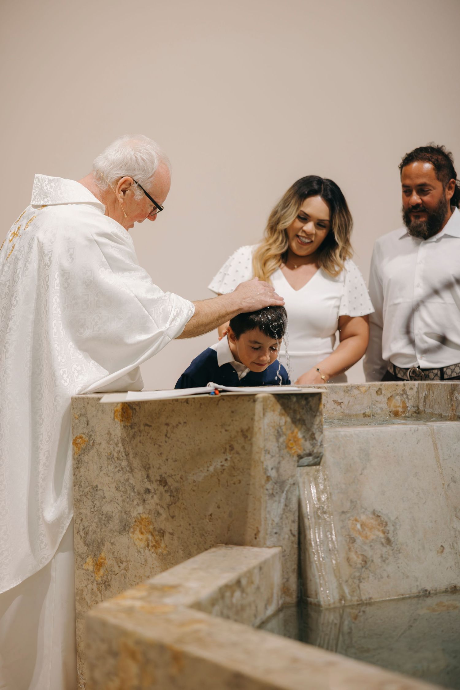Priest baptizing a young boy at a baptismal font with family present.