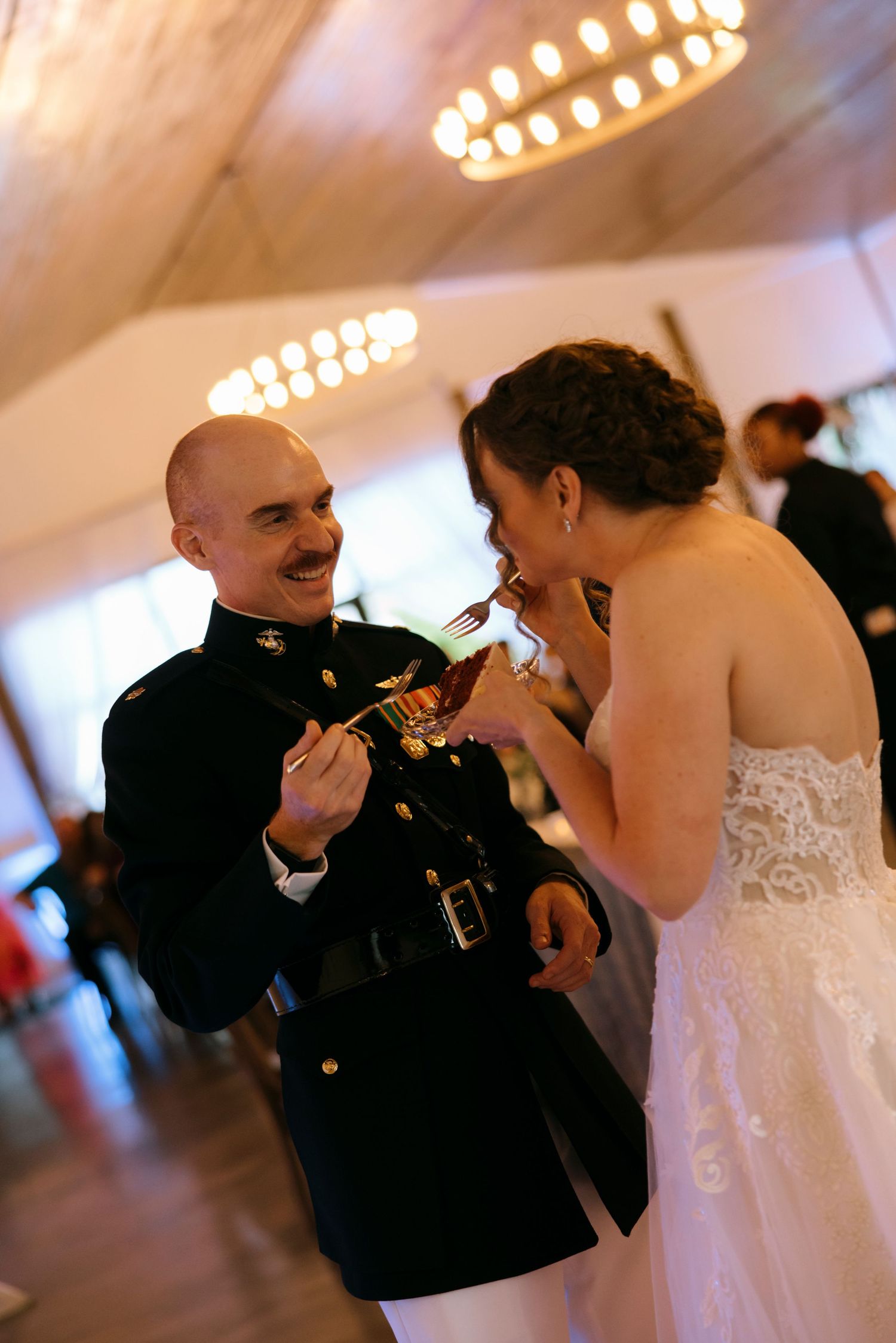 Military groom in dress uniform feeds wedding cake to bride in lace gown at elegant reception venue.