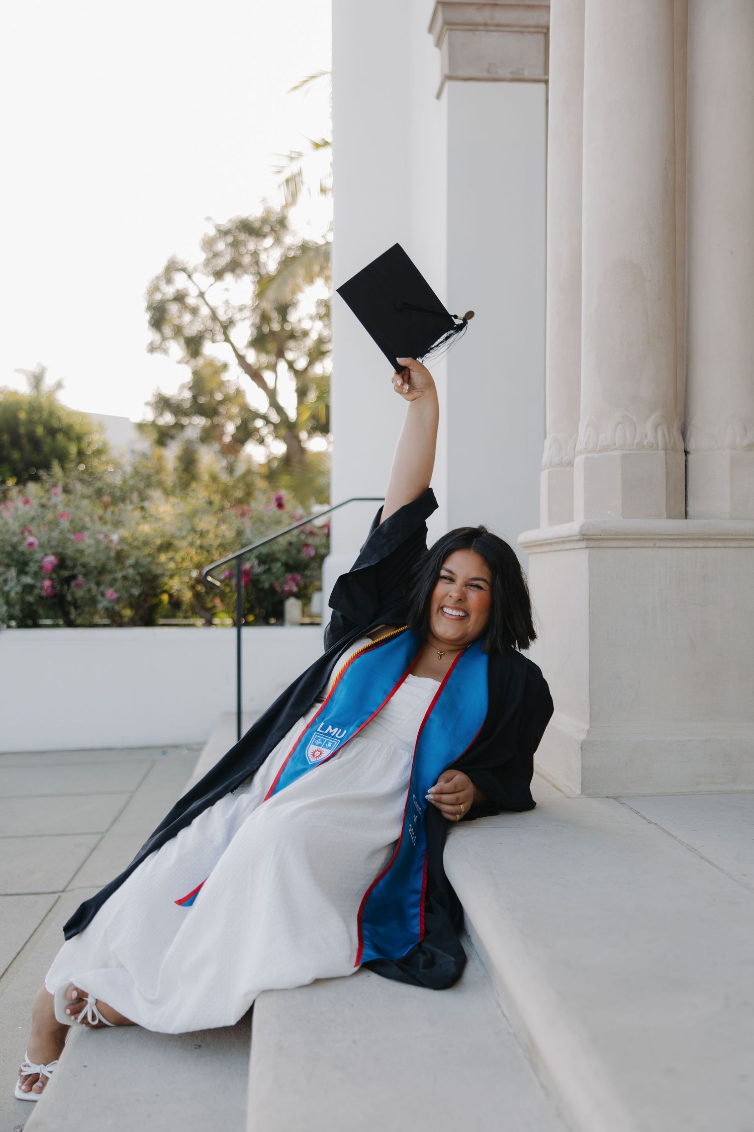 A joyful graduate in black gown and blue stole raises her cap while sitting on steps outdoors.
