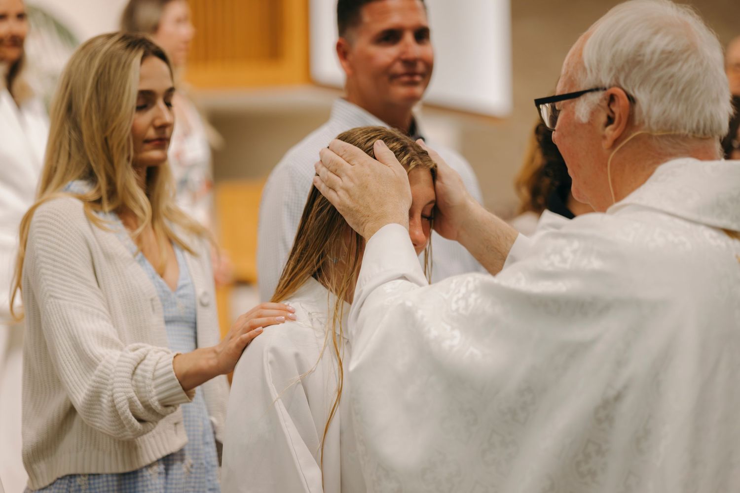 Priest performing a religious blessing ceremony on a young girl in white, with a woman placing her hand on her shoulder.