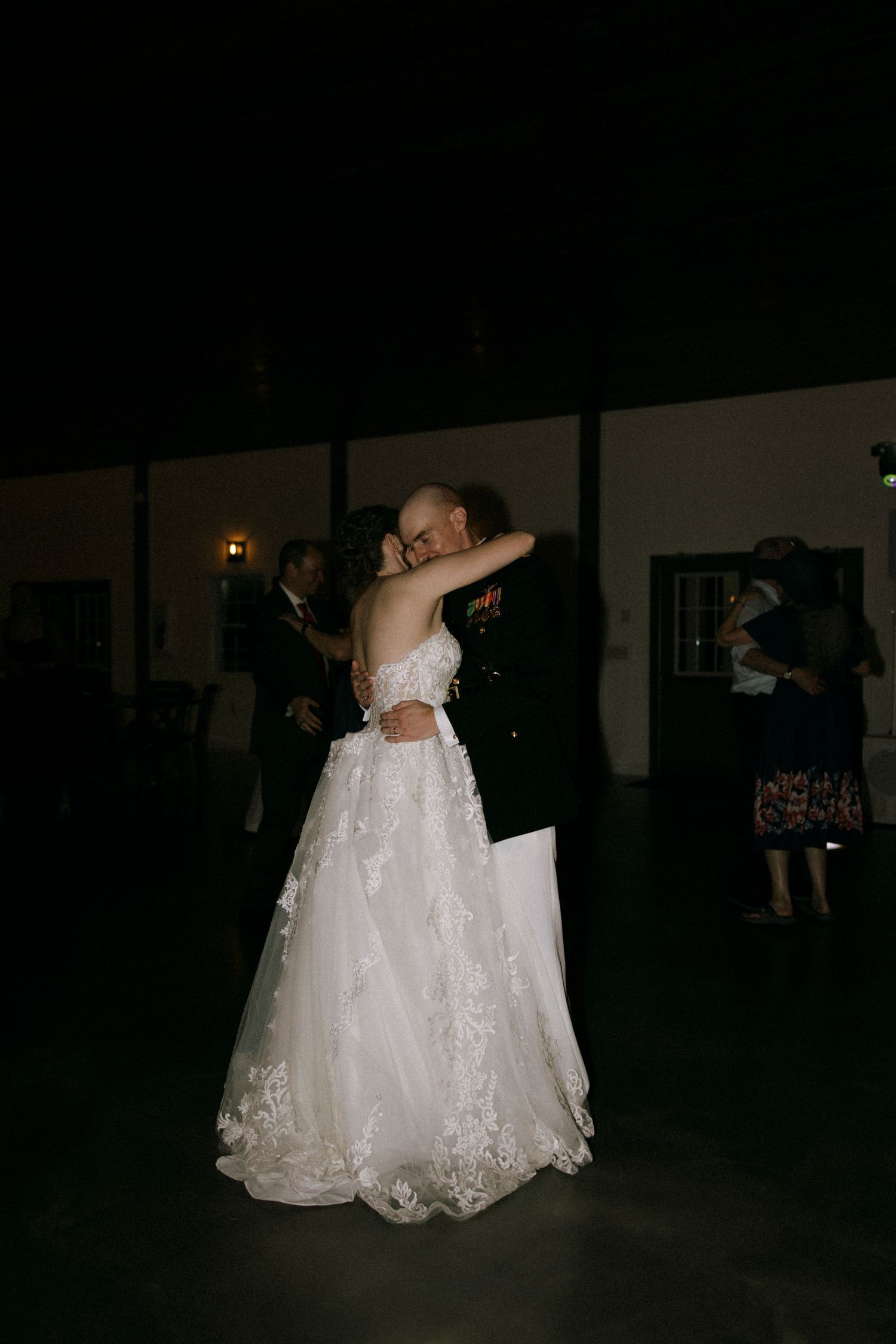 Bride and groom share first dance, bride in lace gown, groom in military dress uniform, dimly lit wedding venue.