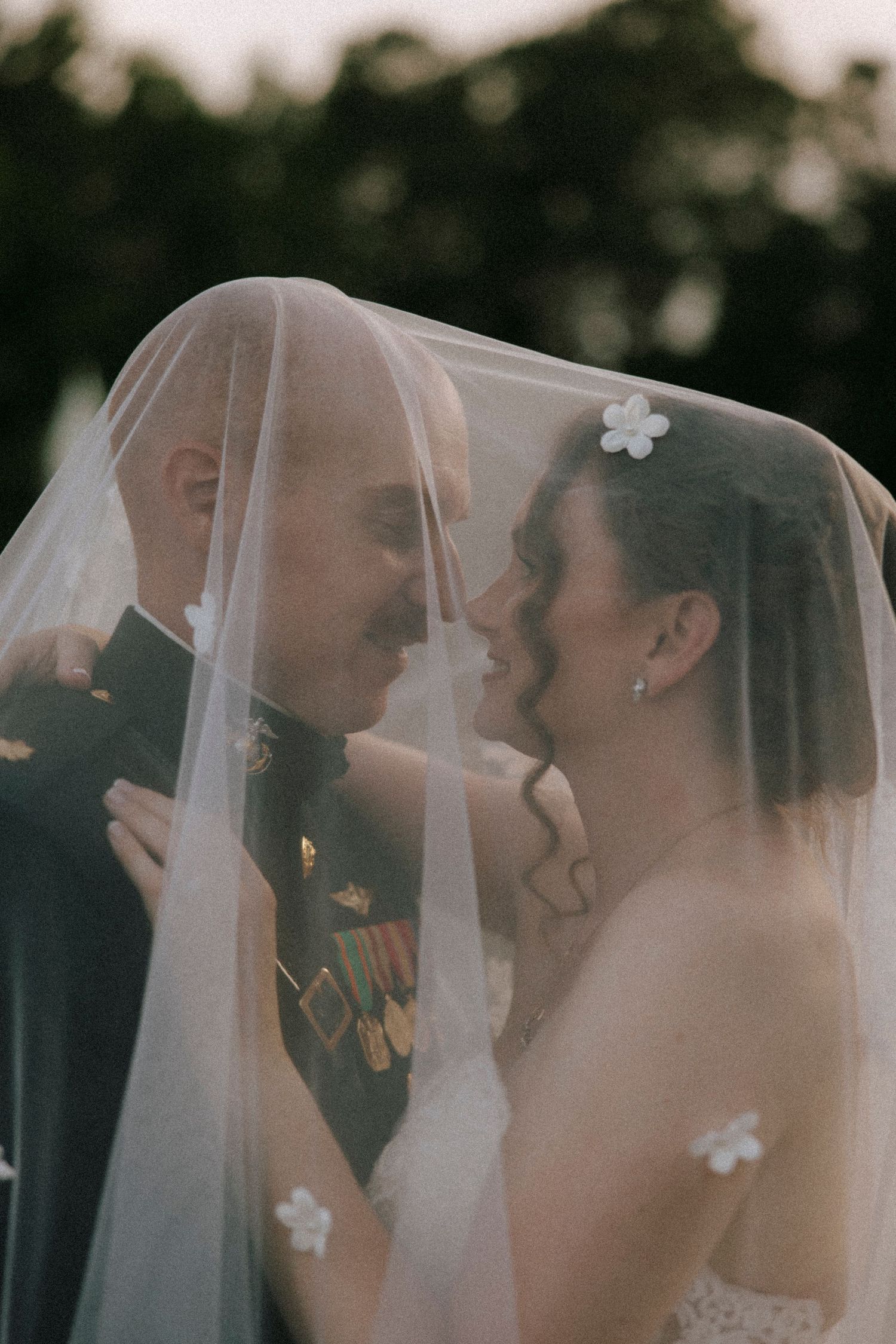 A bride and groom share an intimate moment under a flowing wedding veil, the groom wearing military medals.
