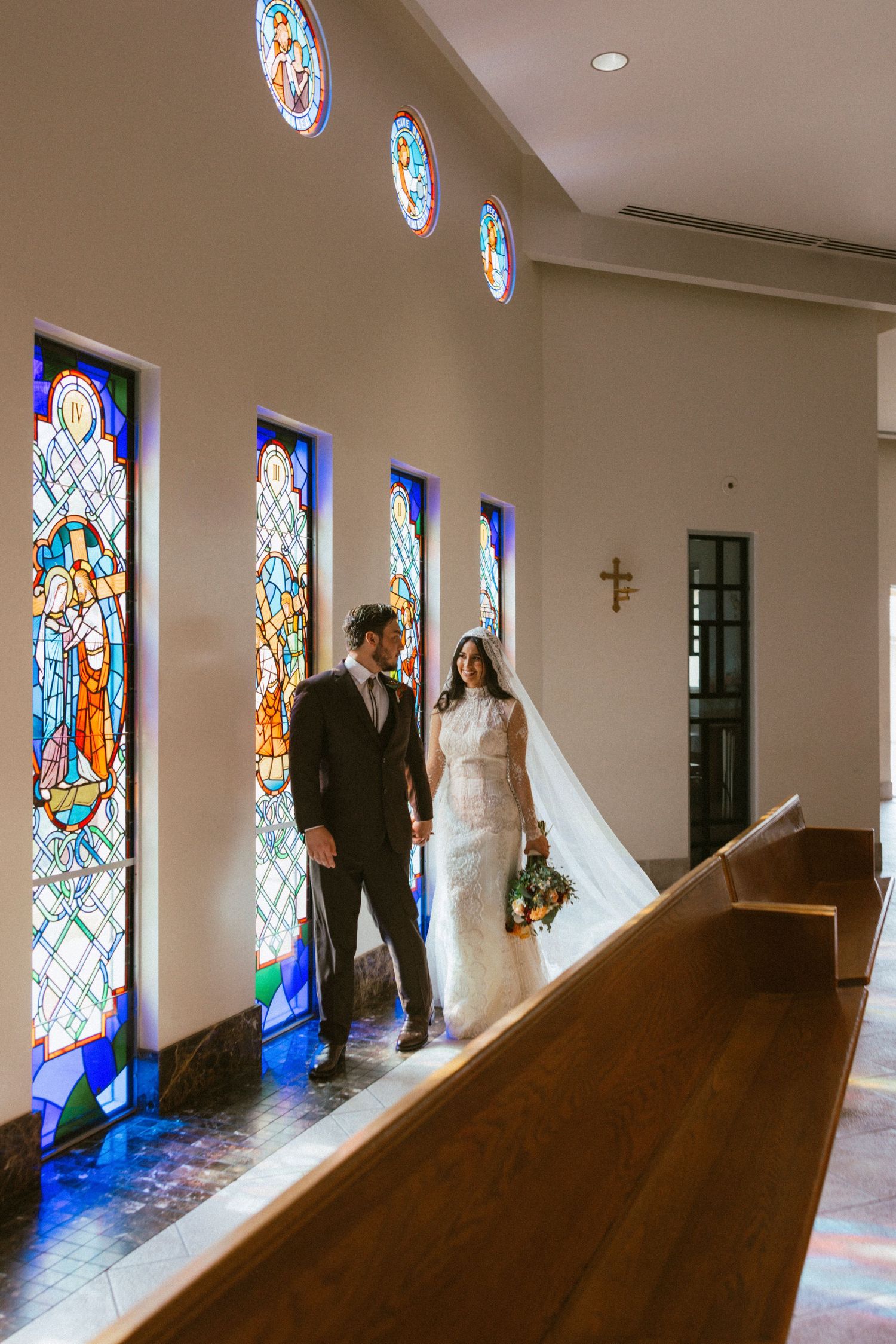 A couple stands between beautiful stained glass windows in a church aisle.