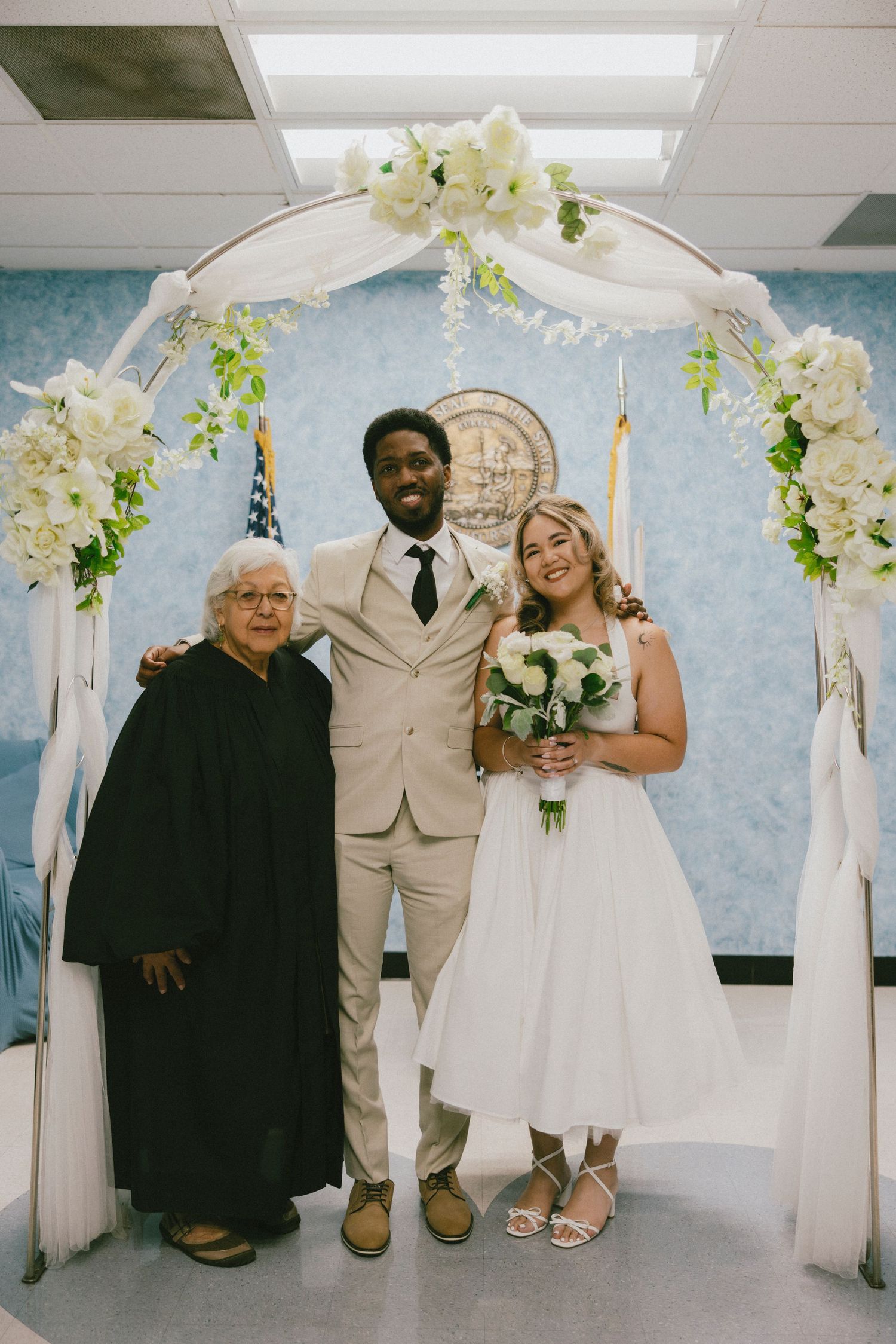 Wedding couple poses with family member under white floral arch at indoor ceremony venue.
