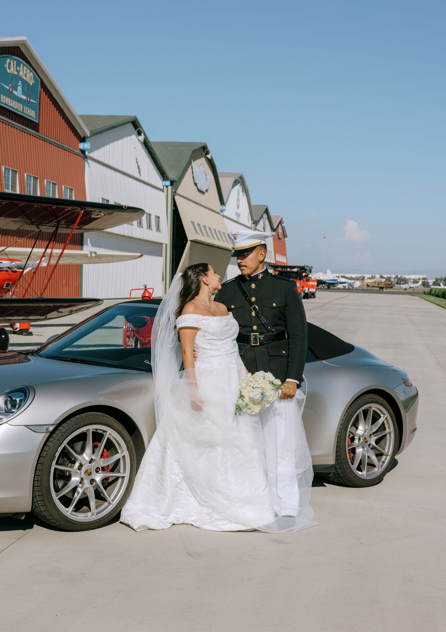 Newlyweds share romantic moments beside luxury sports cars under bright blue sky.