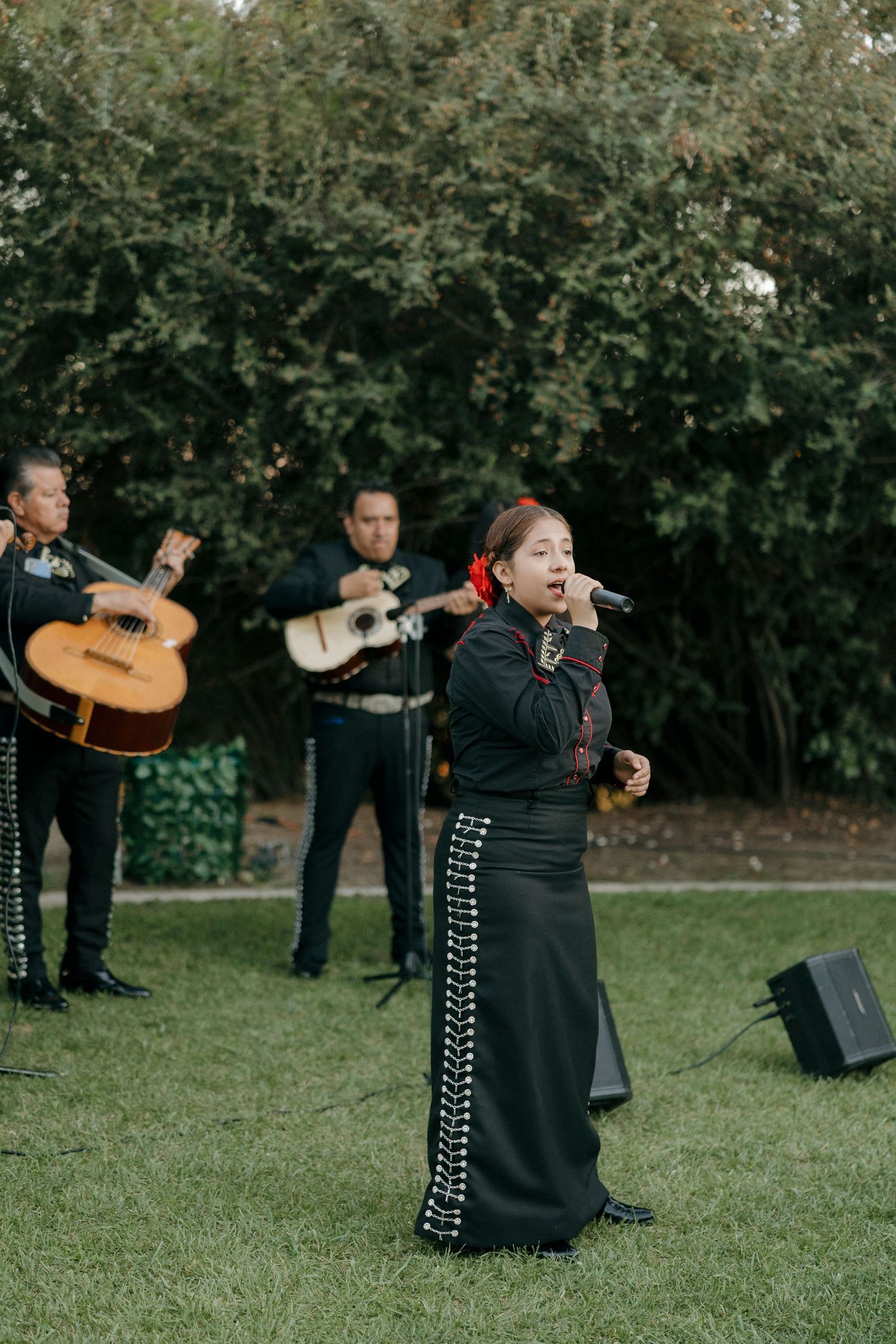 A mariachi singer performs with band members playing guitars in a garden setting.