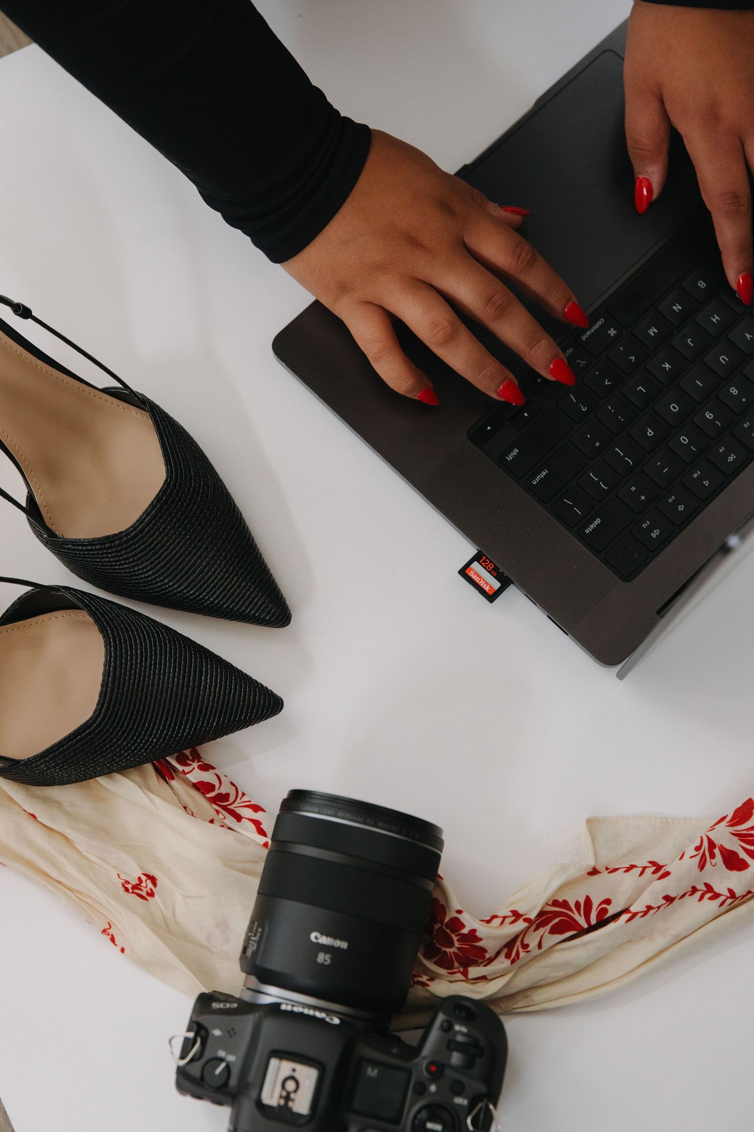 Woman with red nails typing on laptop beside black heels and Canon camera on white desk.