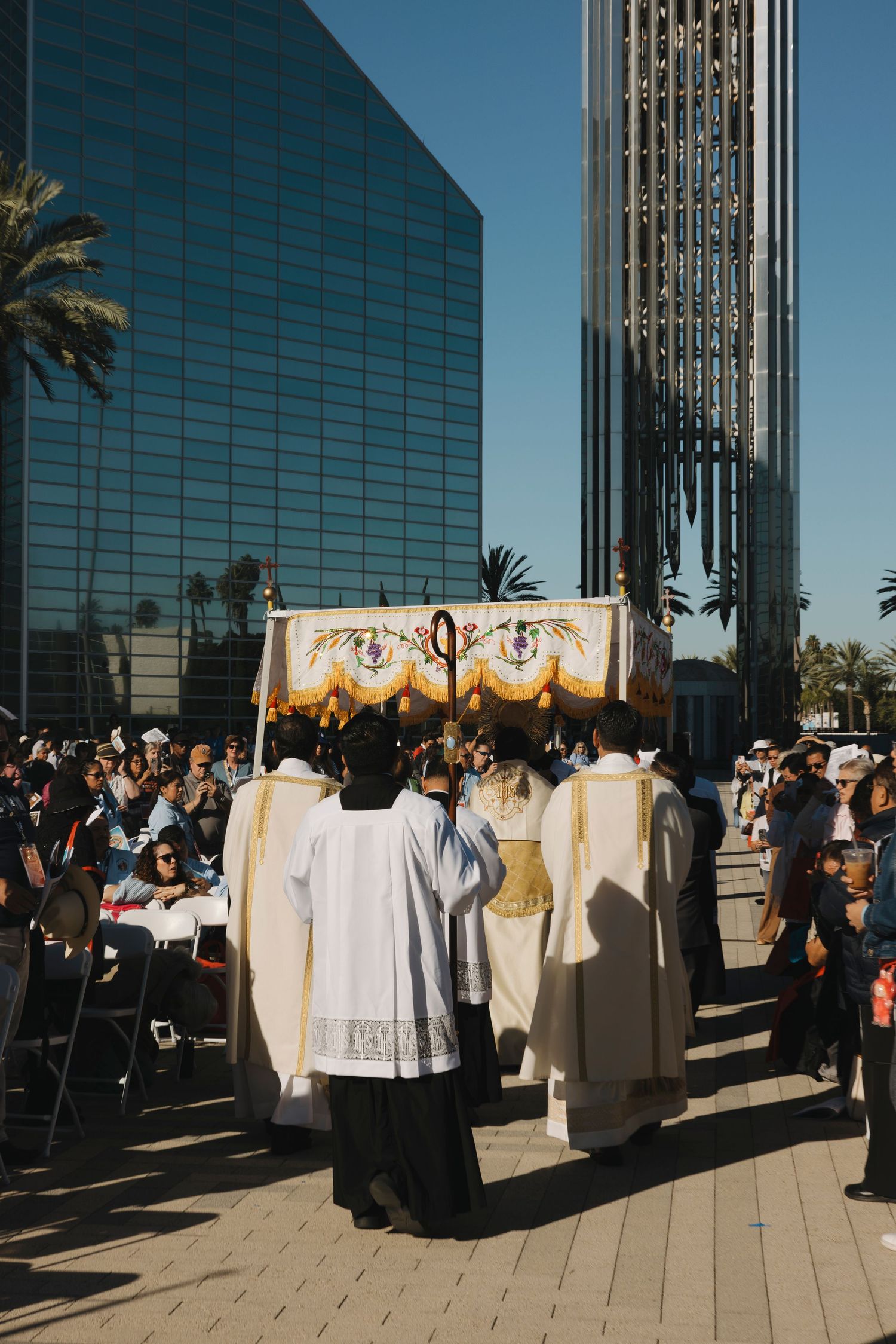 Catholic clergy carry an ornate canopy during a religious procession outside a modern glass building.
