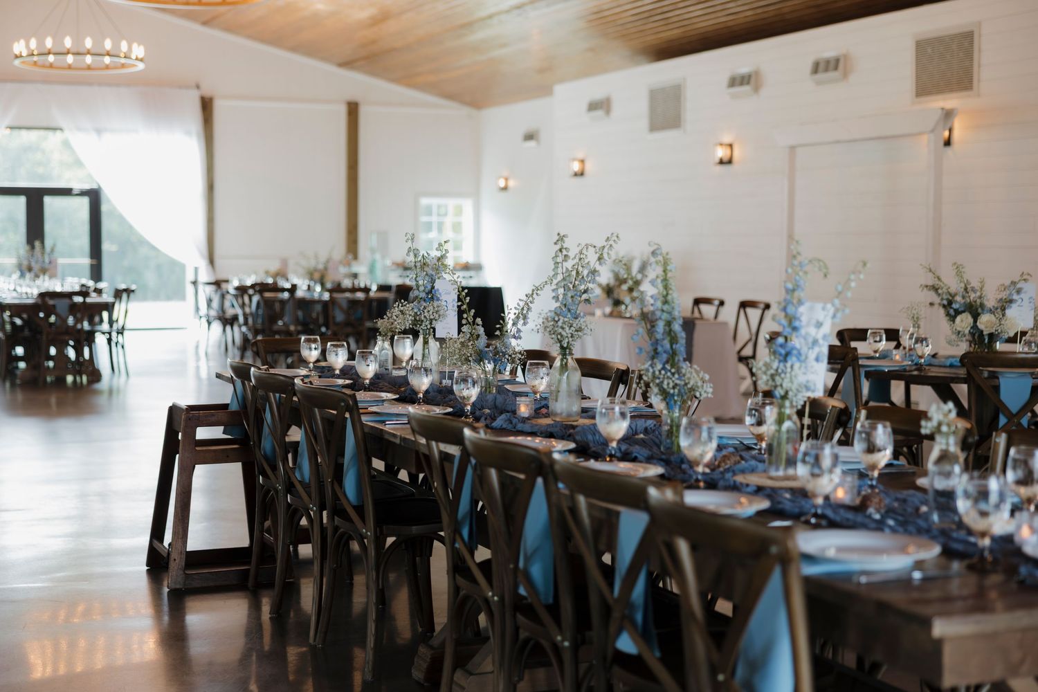 Elegant wedding reception hall with long tables, dark wood chairs, blue napkins, and floral centerpieces in a bright white venue.