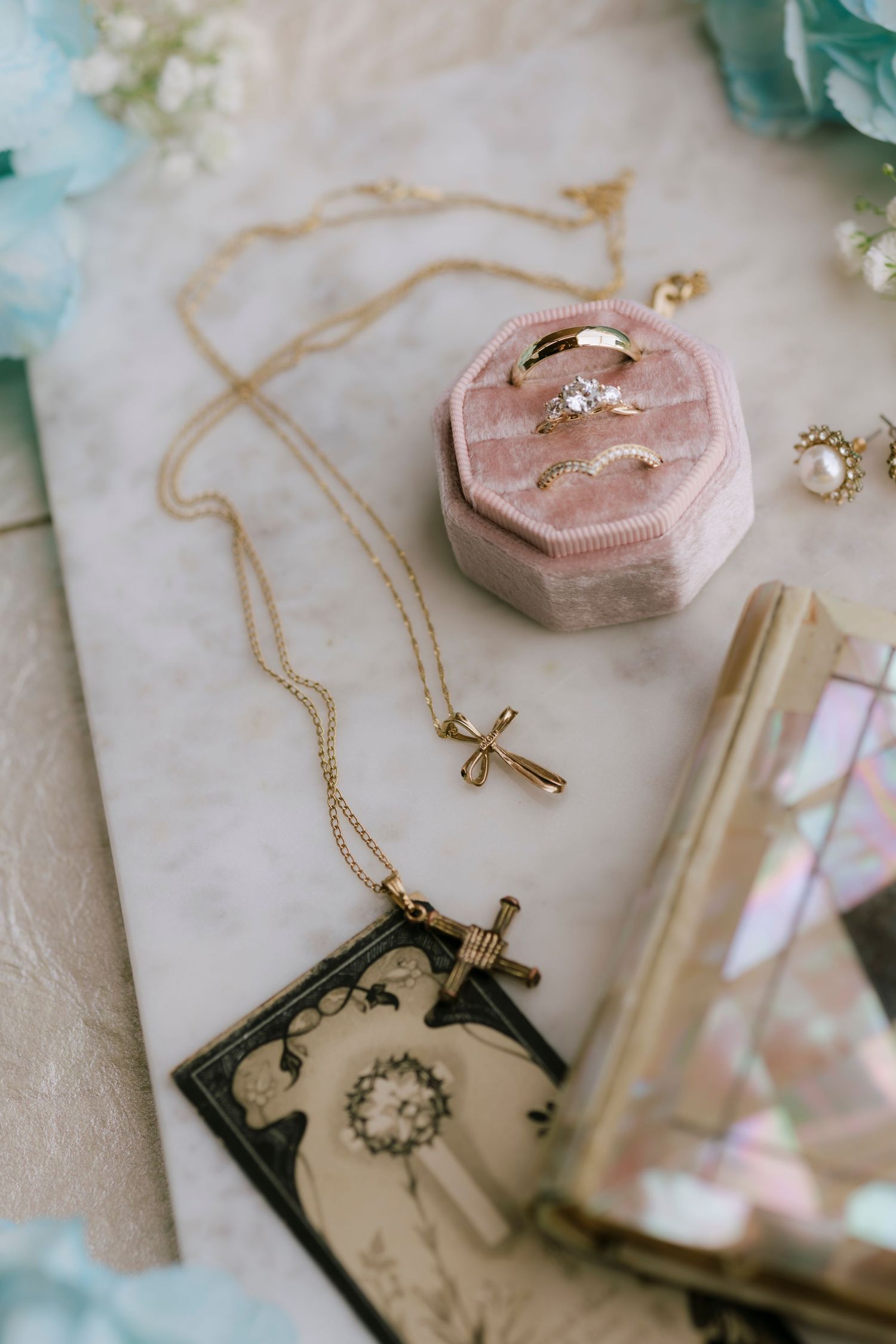 Gold cross necklaces and wedding rings in a pink velvet box on marble, surrounded by pearl earrings and blue flowers.