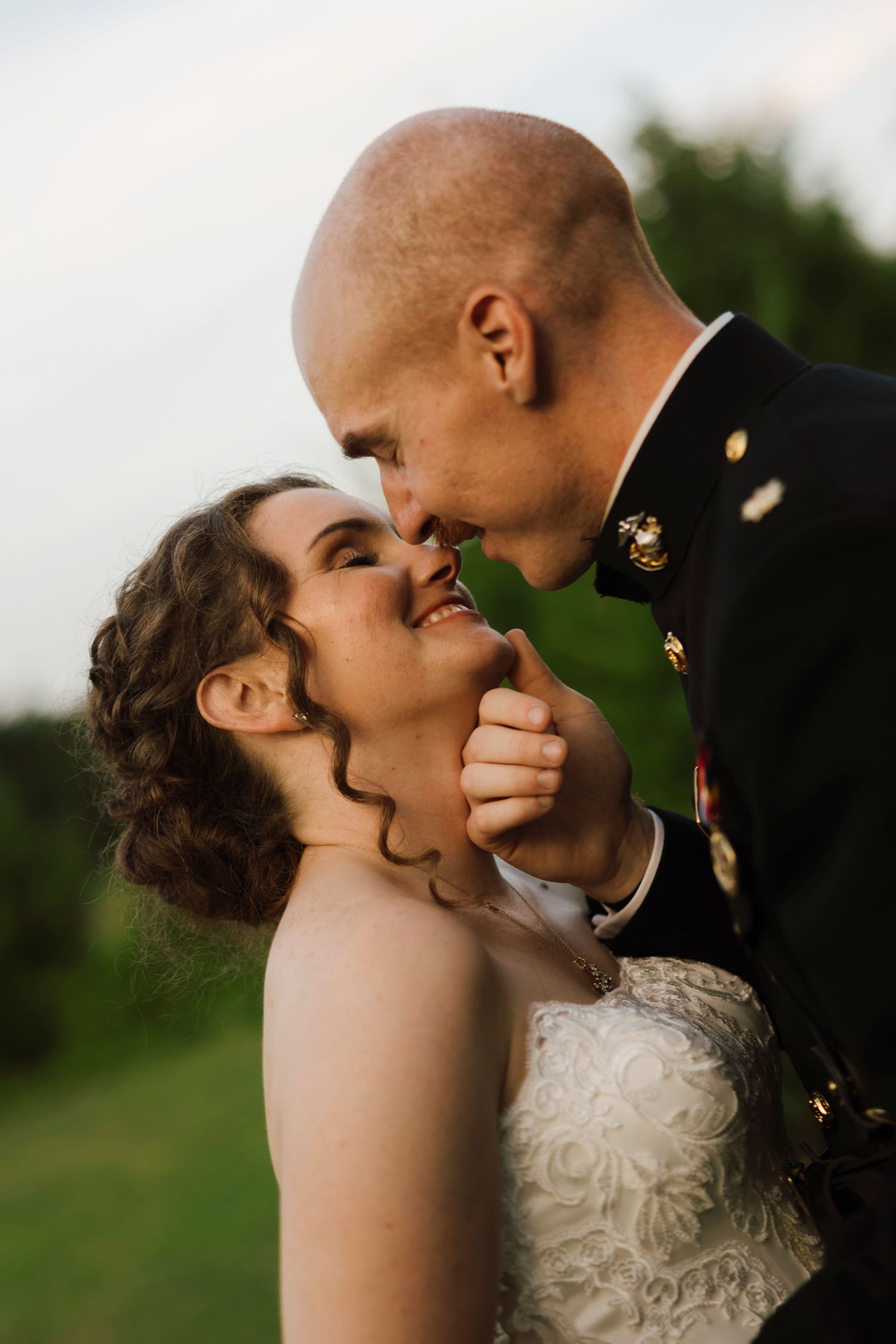 A bride in white lace and groom in military dress uniform share a romantic moment outdoors at their wedding.