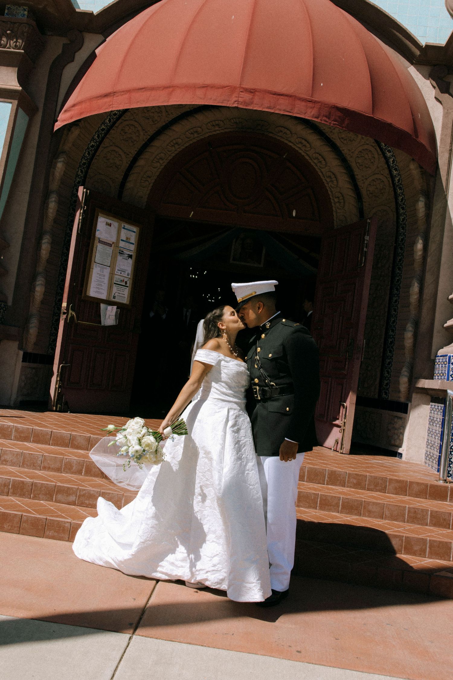 Wedding party members in formal attire stand in a row outside a brick archway entrance.