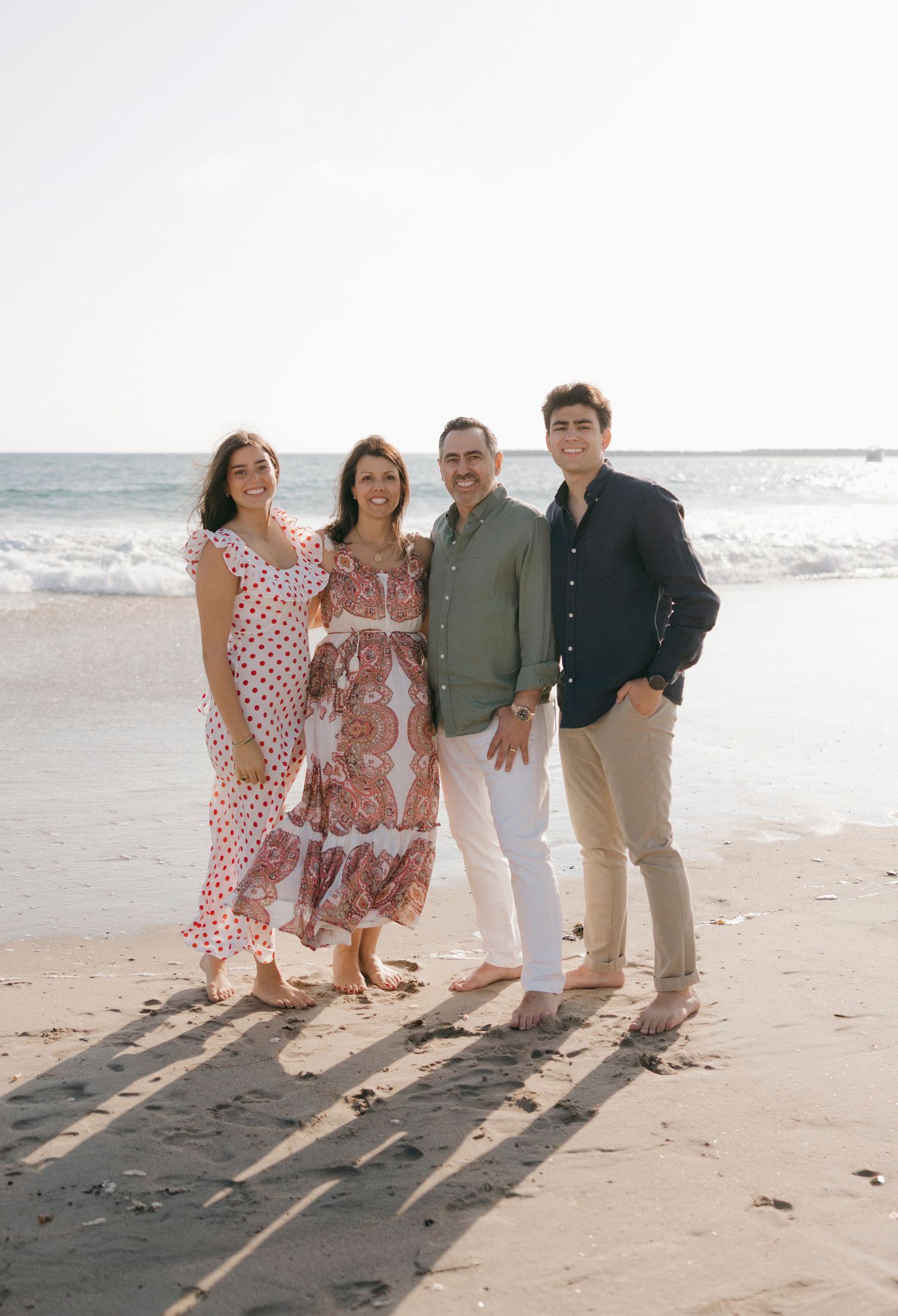Family of four smiling together on a sunny beach, wearing casual summer outfits with ocean waves in the background.
