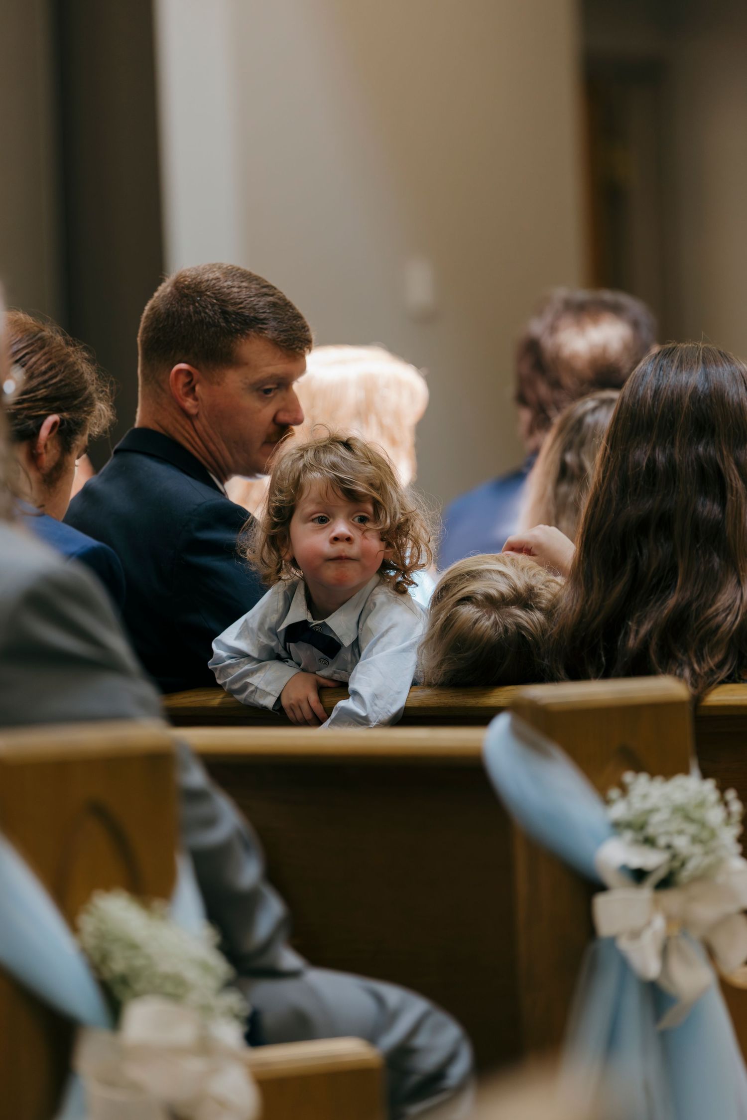 A young child sits on an adult's lap during a wedding ceremony in a church pew decorated with blue ribbons and white flowers.