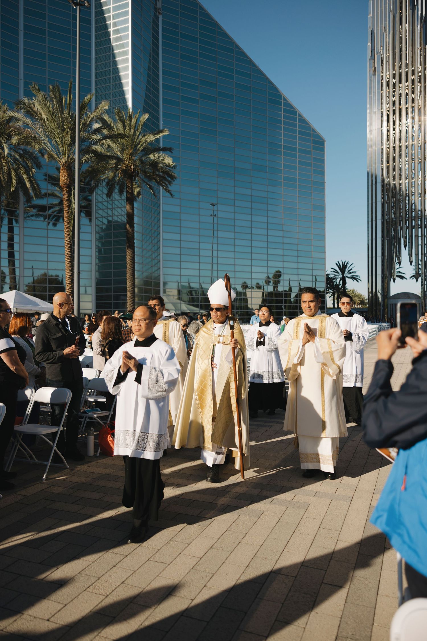 Religious ceremony participants gather outside a modern glass building with palm trees.
