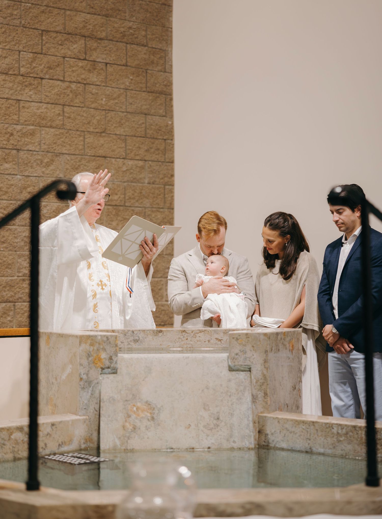Priest performing a baptism ceremony with parents holding infant at stone baptismal font in modern church.