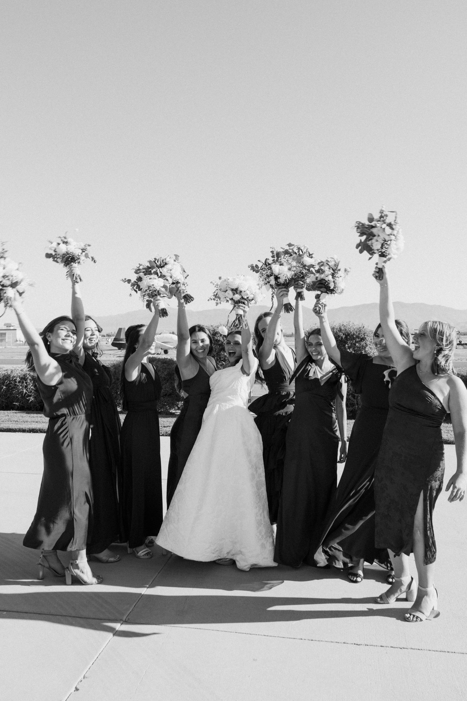 Black and white photo of wedding party raising bouquets in celebration outdoors.