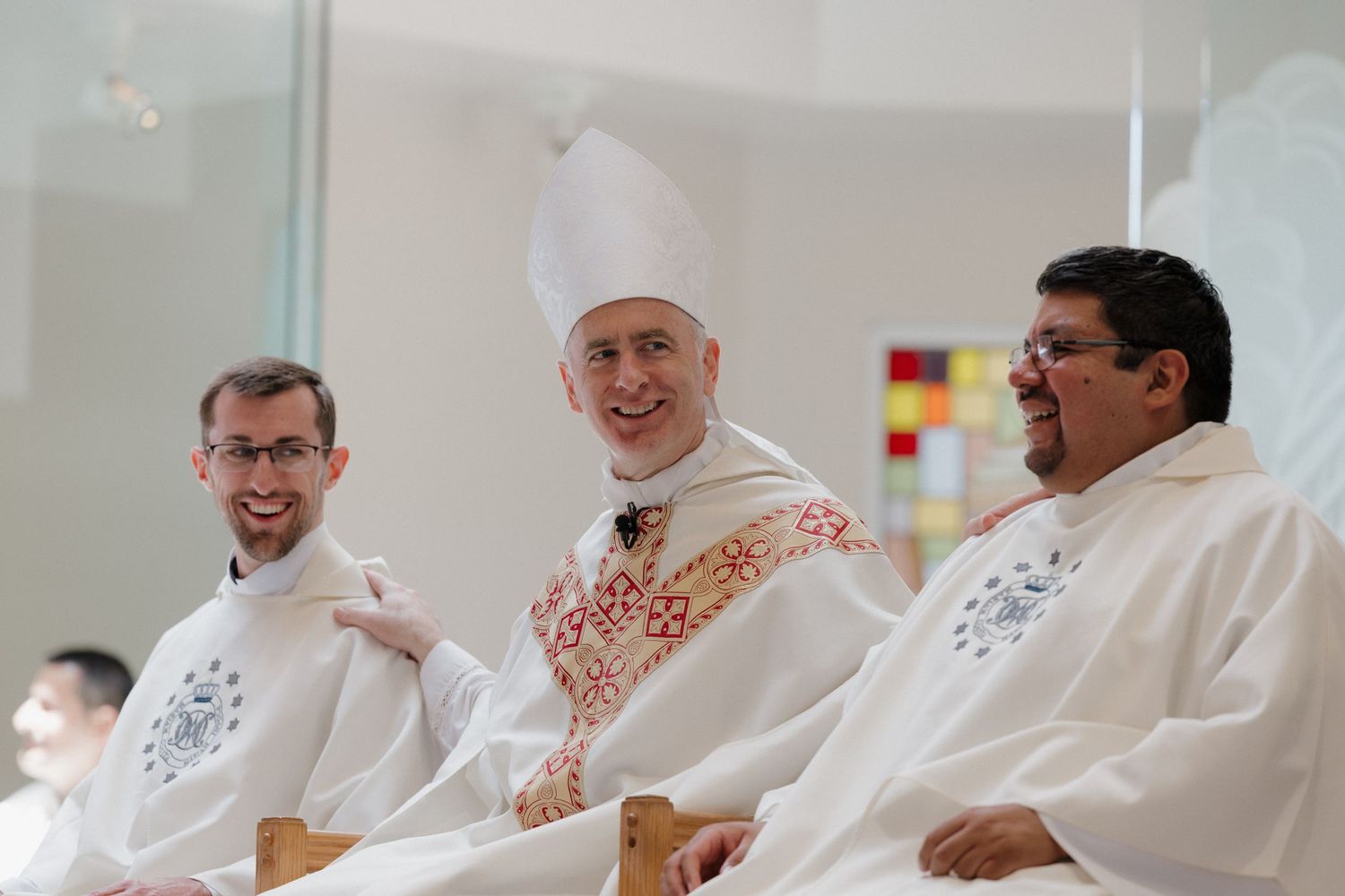 A group of clergy members in white robes seated together and smiling during a religious gathering.