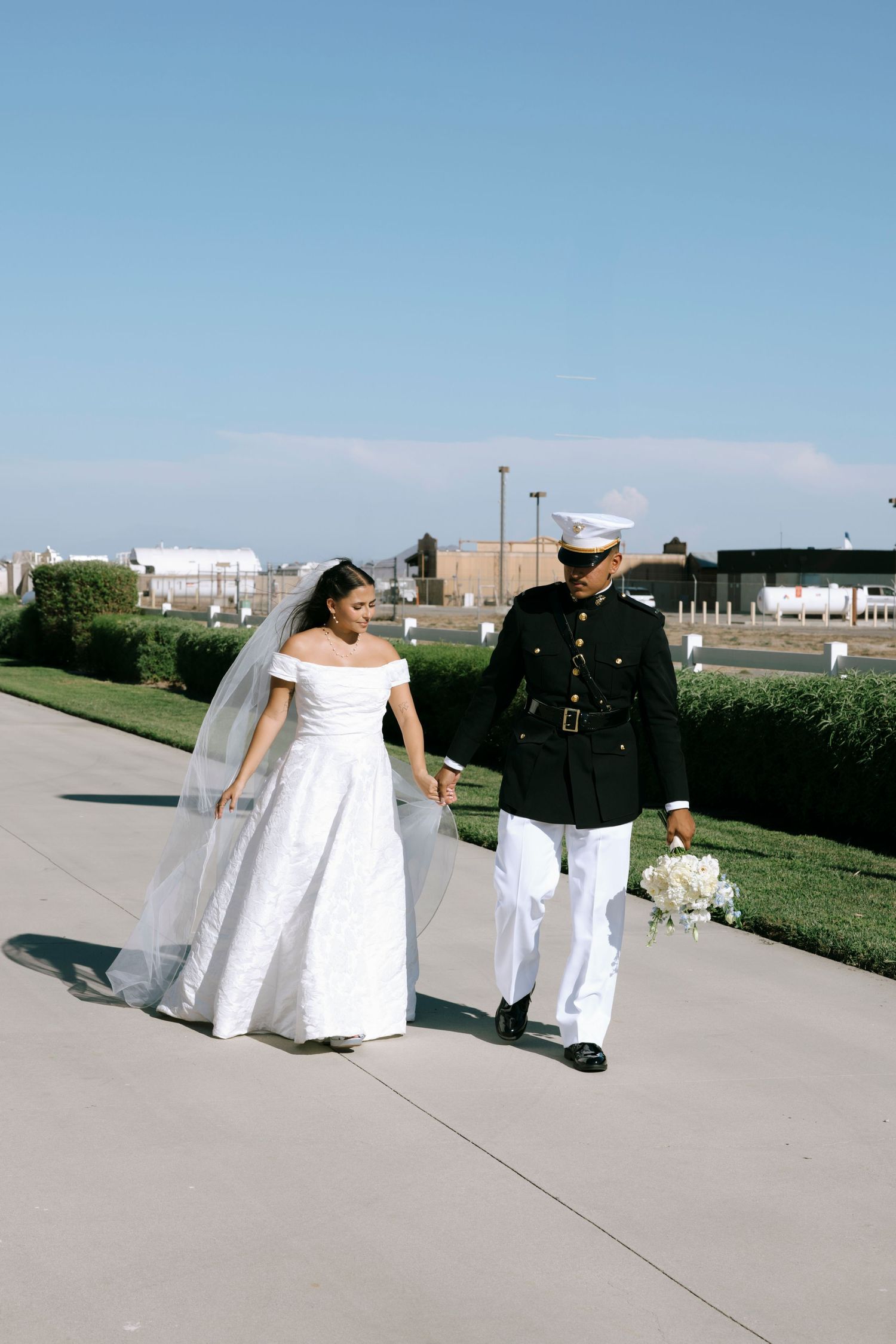 A military service member and bride walk together on a sunny day with buildings in the background.