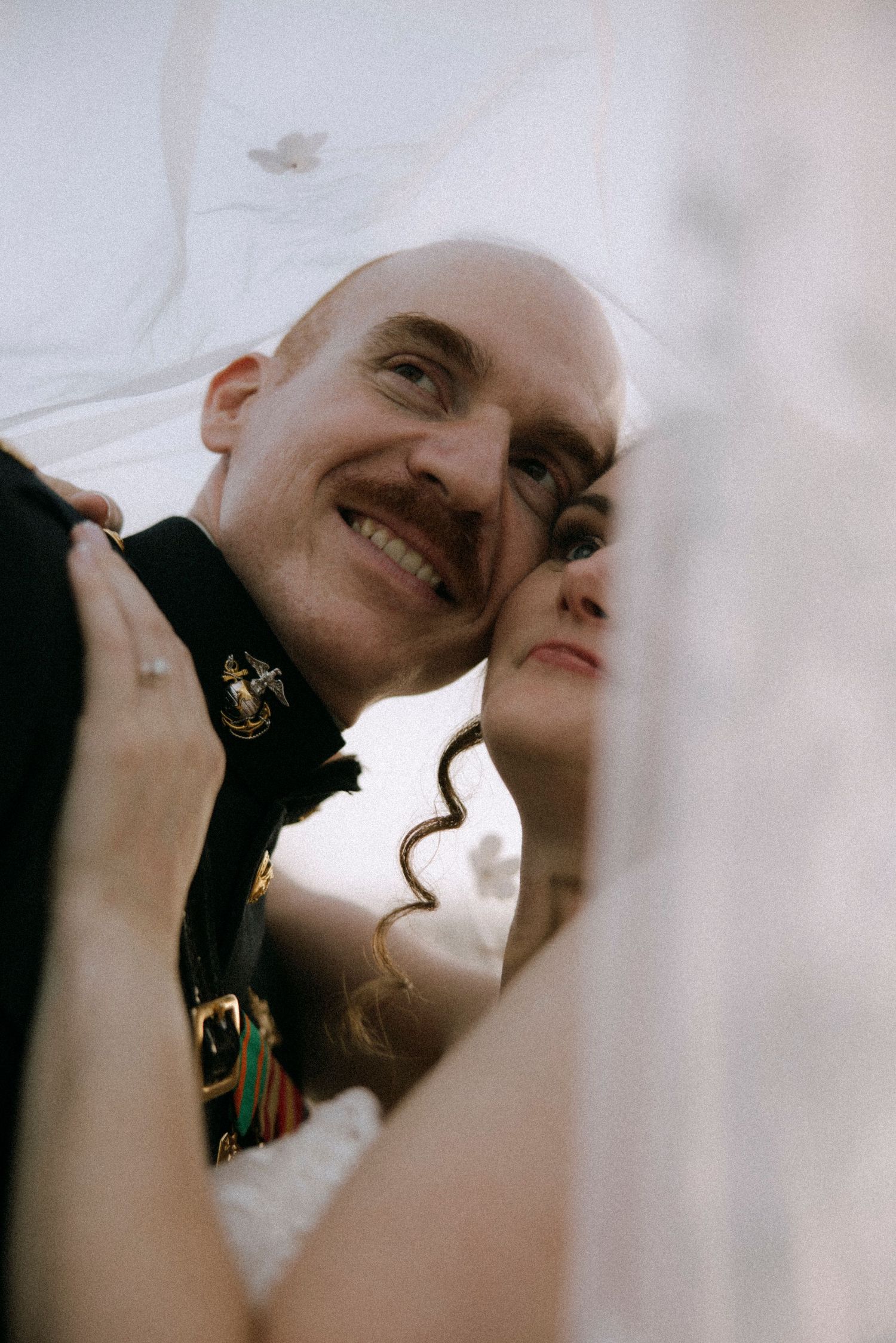 Smiling groom in military dress uniform poses with bride under wedding veil in romantic close-up portrait.