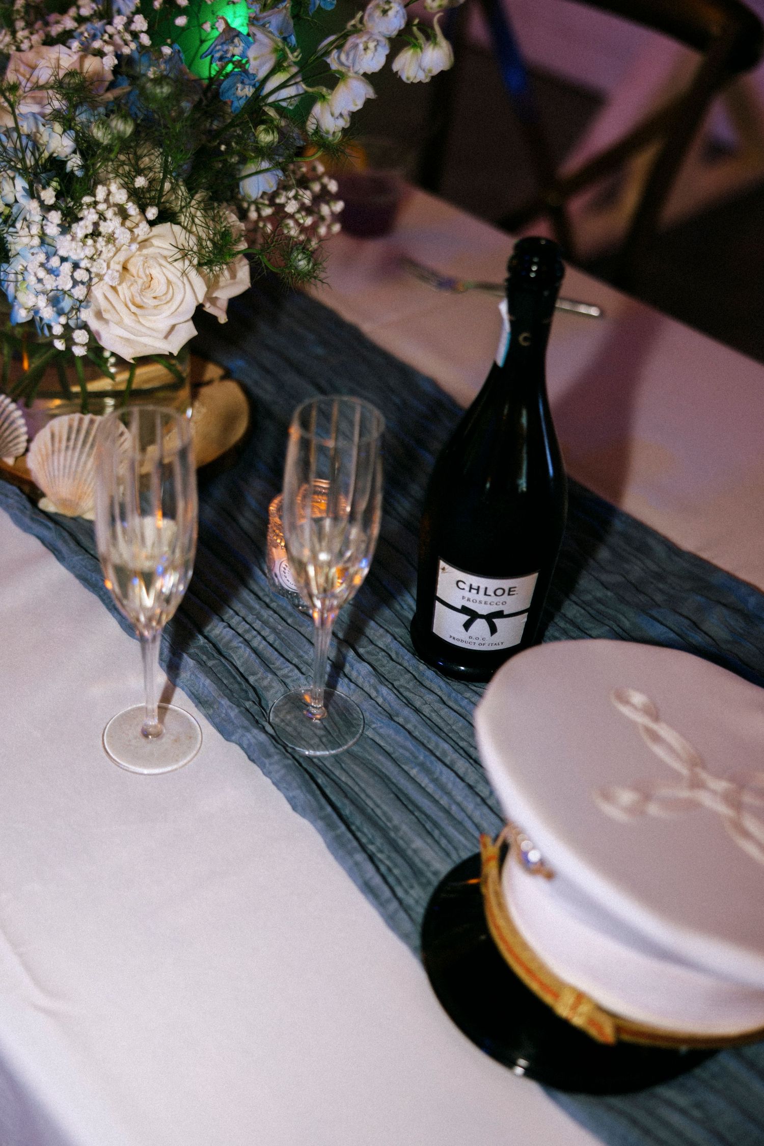 Elegant wedding table with Chloe sparkling wine, crystal champagne flutes, white roses, and a nautical captain's hat centerpiece.