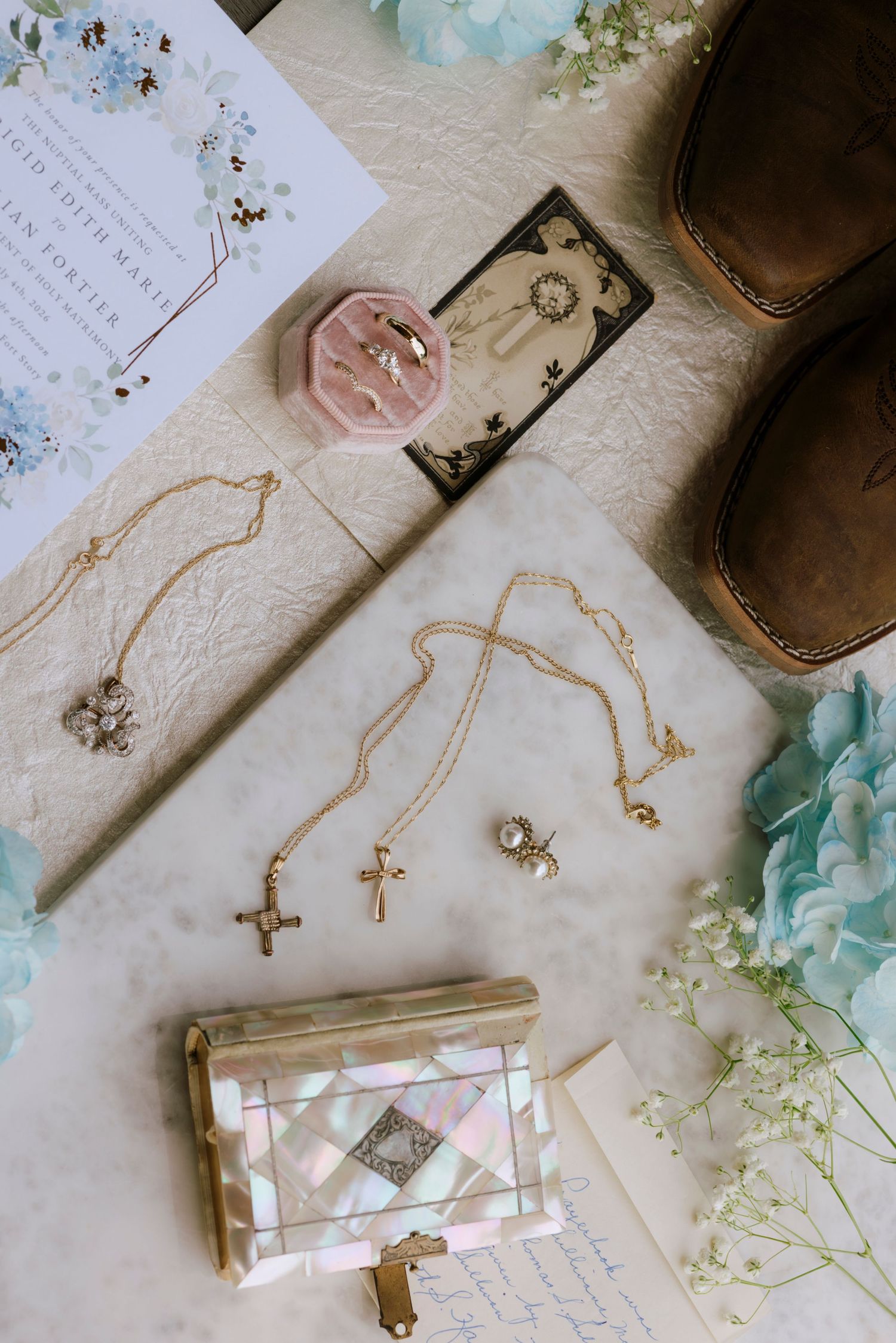 Gold cross necklaces, pearl clutch, ring box, brown boots, and floral wedding stationery arranged on marble surface.