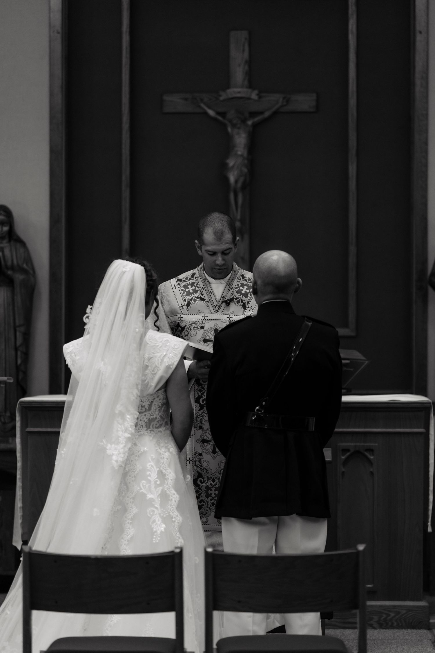 Black and white photo of a bride and groom exchanging vows before a priest at a Catholic church altar with crucifix.