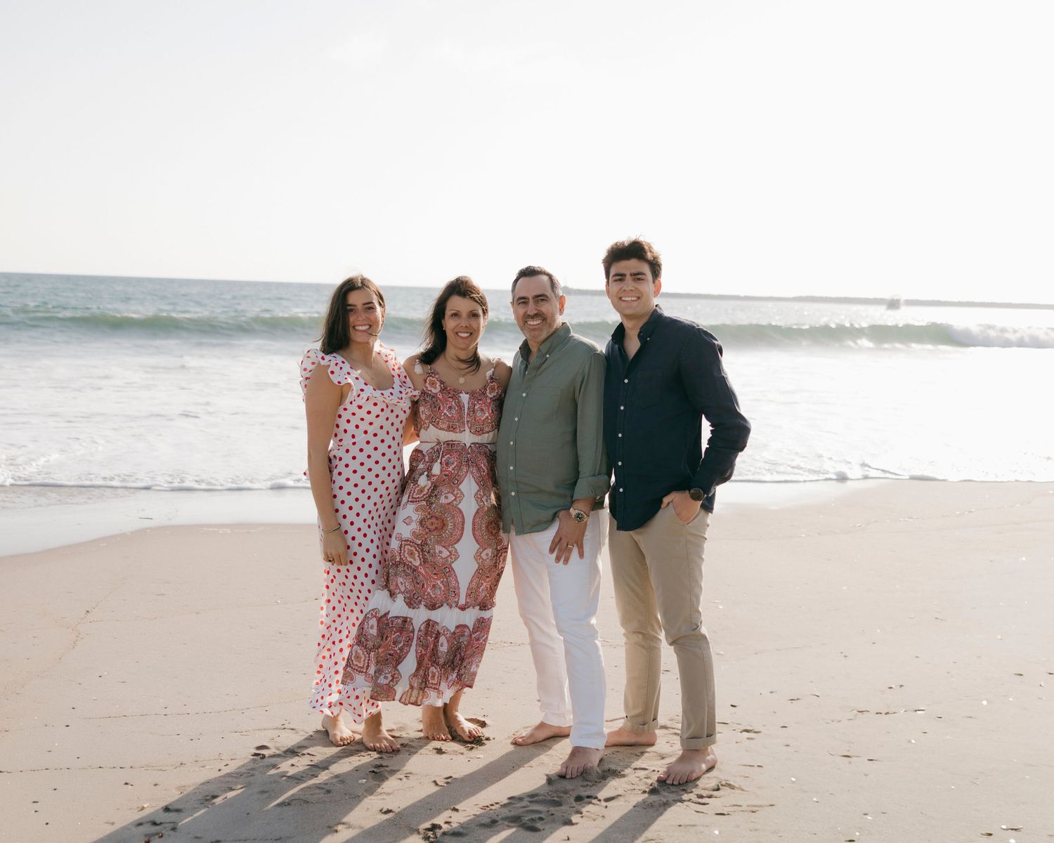 Family of four smiling together on a sunny beach with ocean waves in the background.