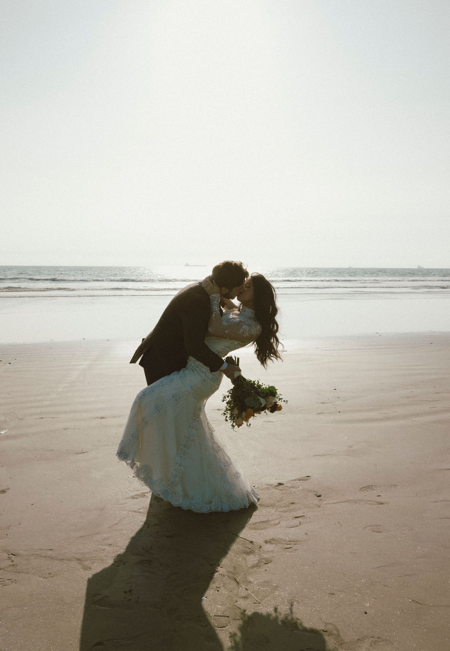 Couple in wedding attire embracing on a sunlit beach with ocean waves in background.