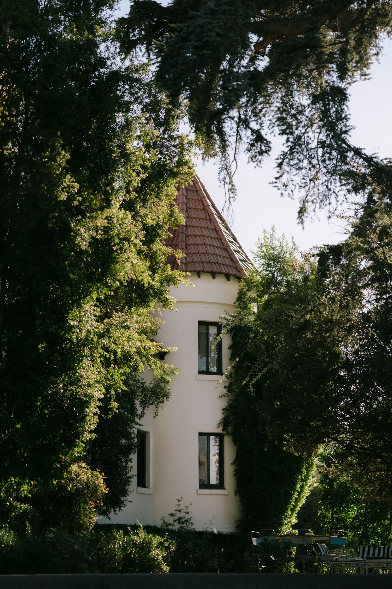 A white tower-style building with a conical roof nestled among lush green trees.